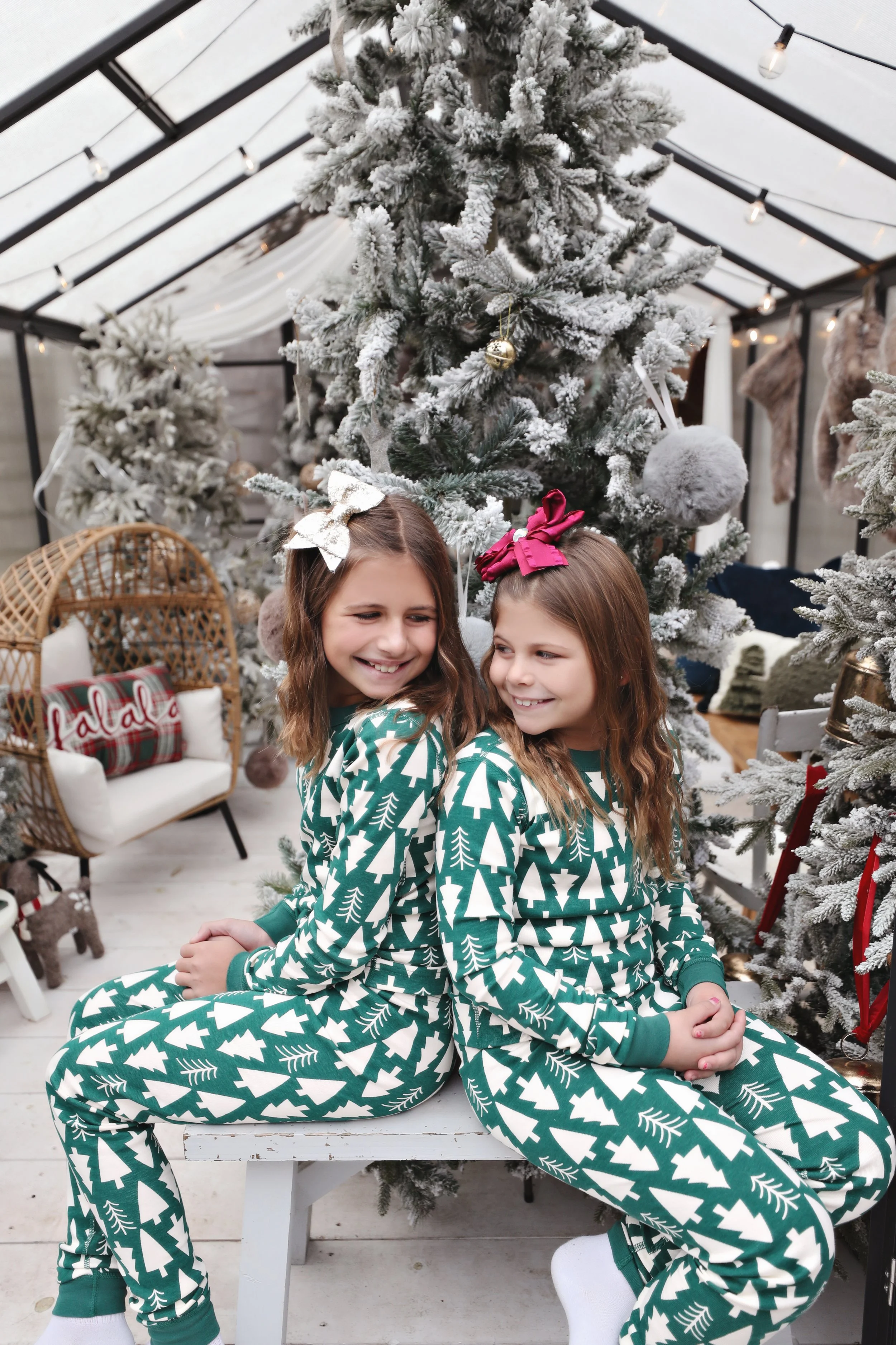 Two young girls sitting back-to-back on a white bench, wearing matching green Christmas pajamas with white trees. They are smiling and looking at each other in front of a decorated Christmas tree inside a well-lit greenhouse or conservatory.