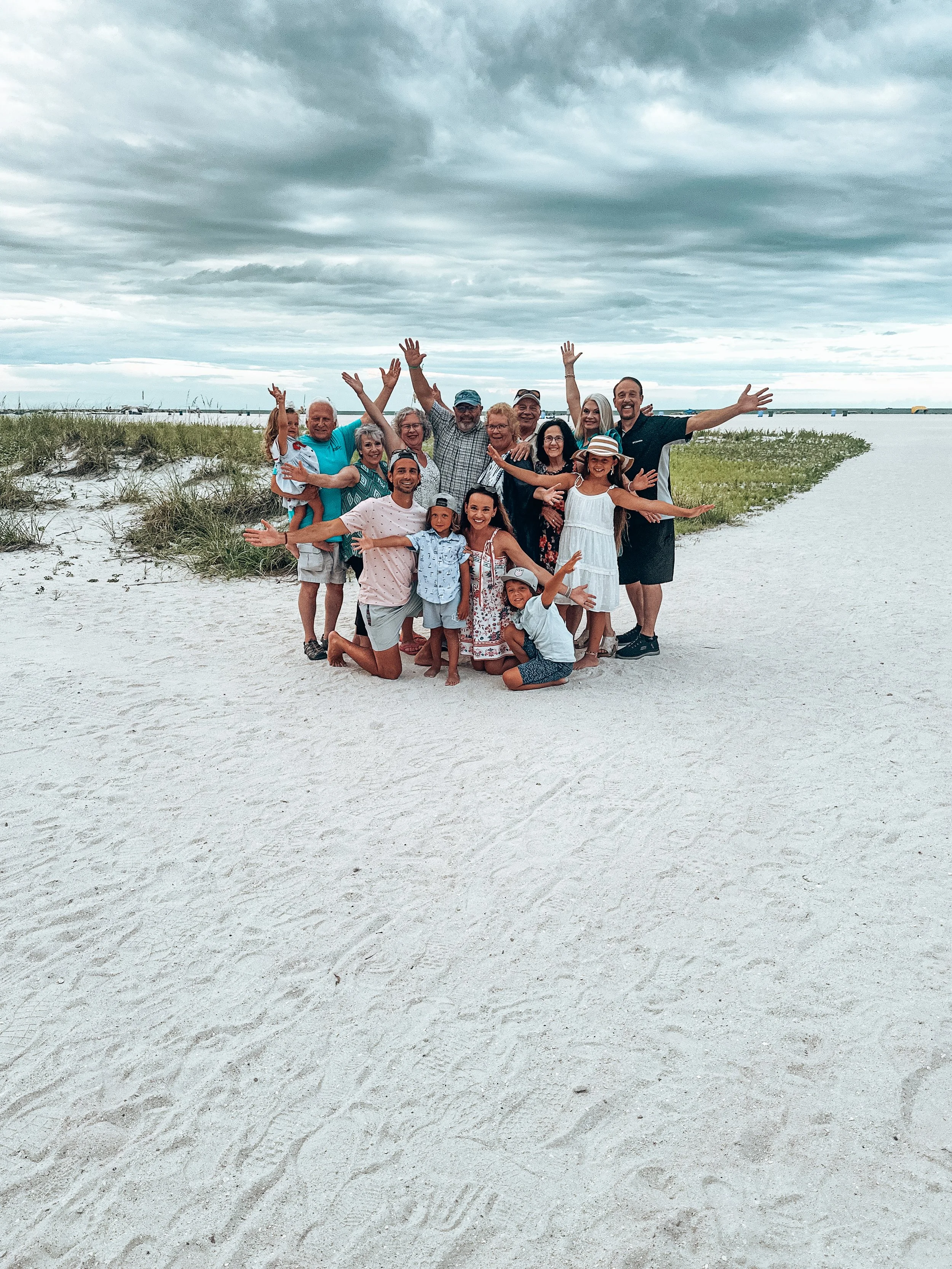 Group of people smiling and posing on a sandy beach with water and cloudy sky in the background.