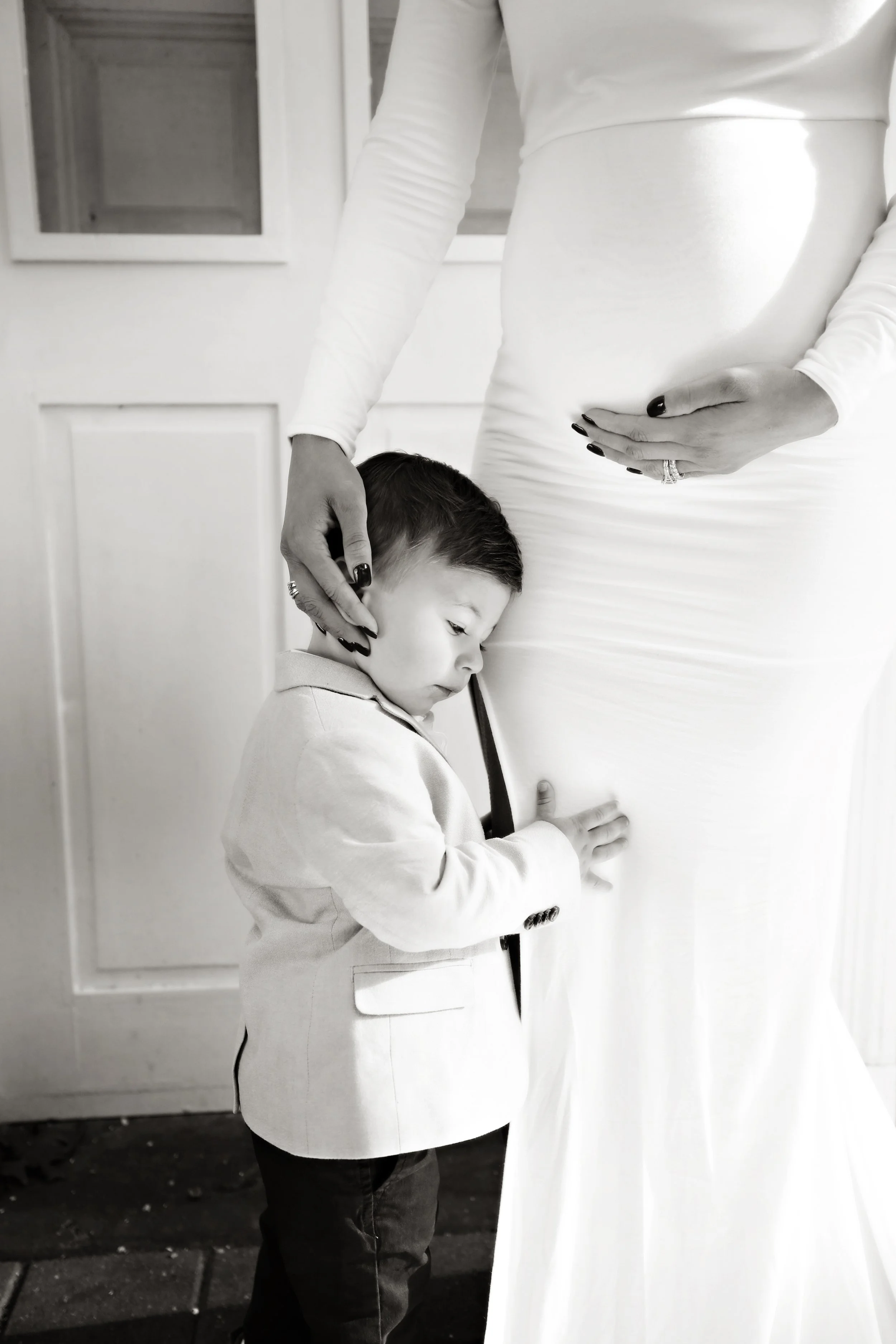 A young boy with his head resting against a woman’s pregnant belly, who is gently touching him with one hand while holding her belly with the other.