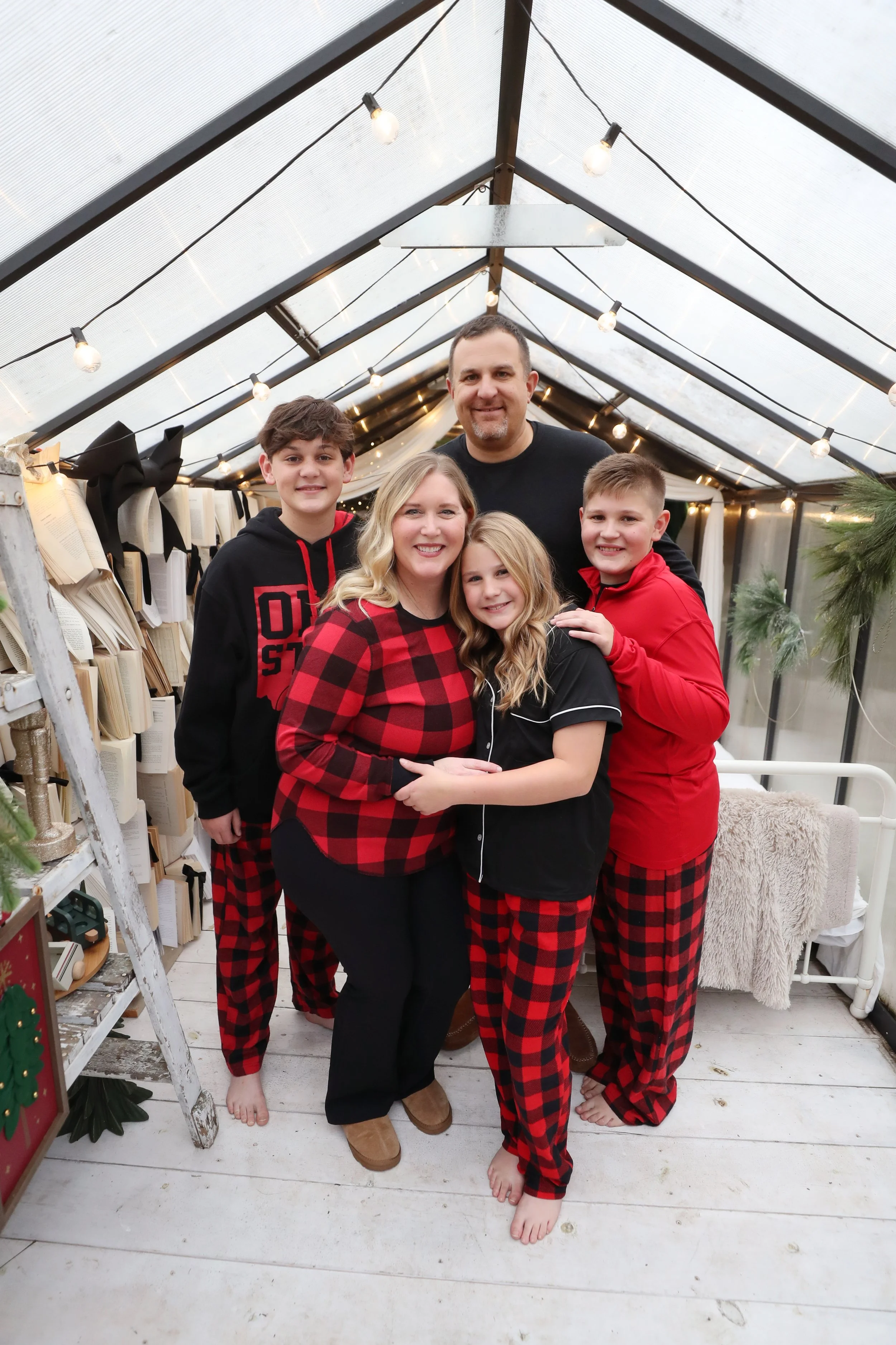 Family in Christmas pajamas inside a greenhouse decorated for the holidays, smiling at the camera.