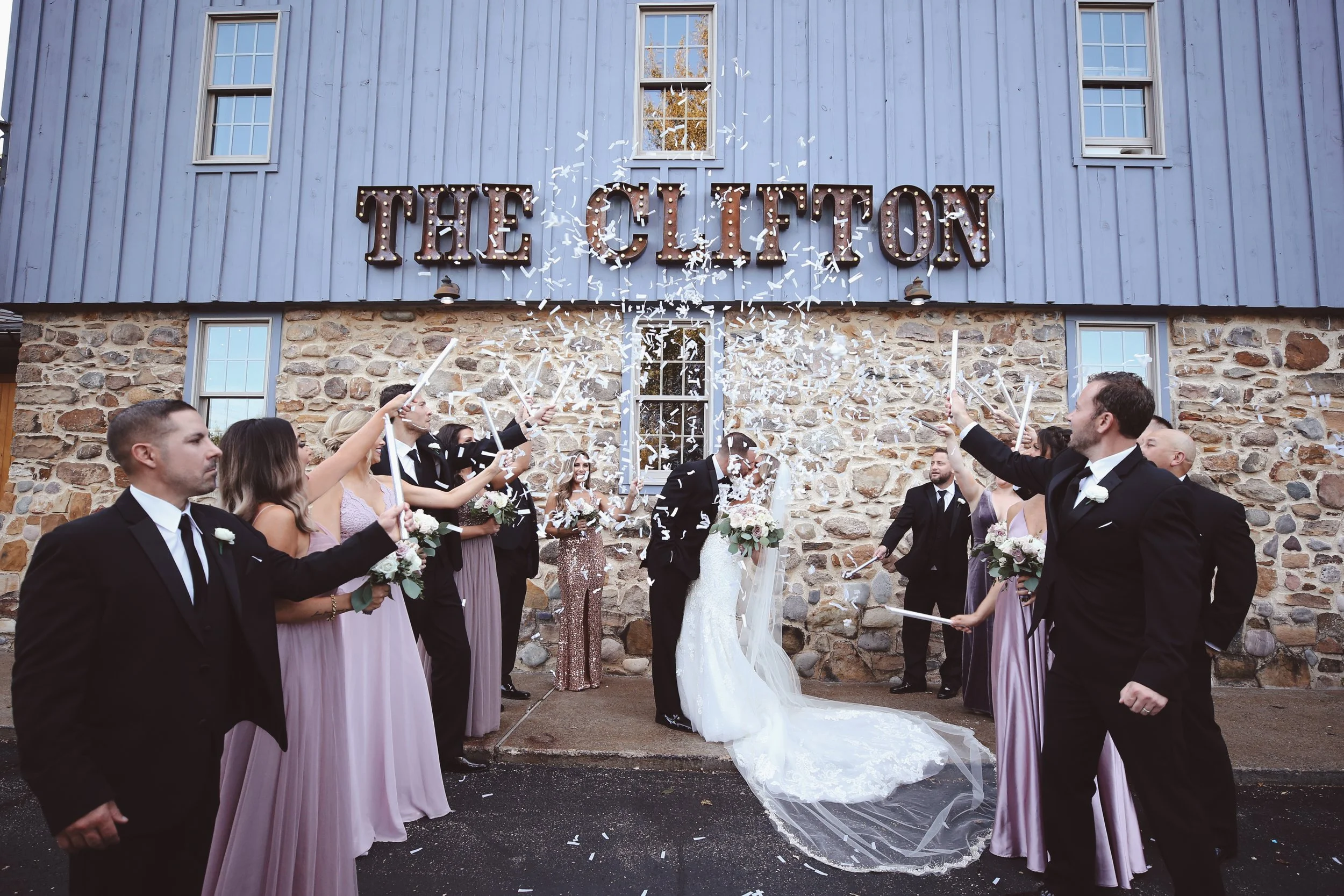 Wedding celebration outside The Clinton building with a bride and groom kissing, surrounded by guests throwing confetti.