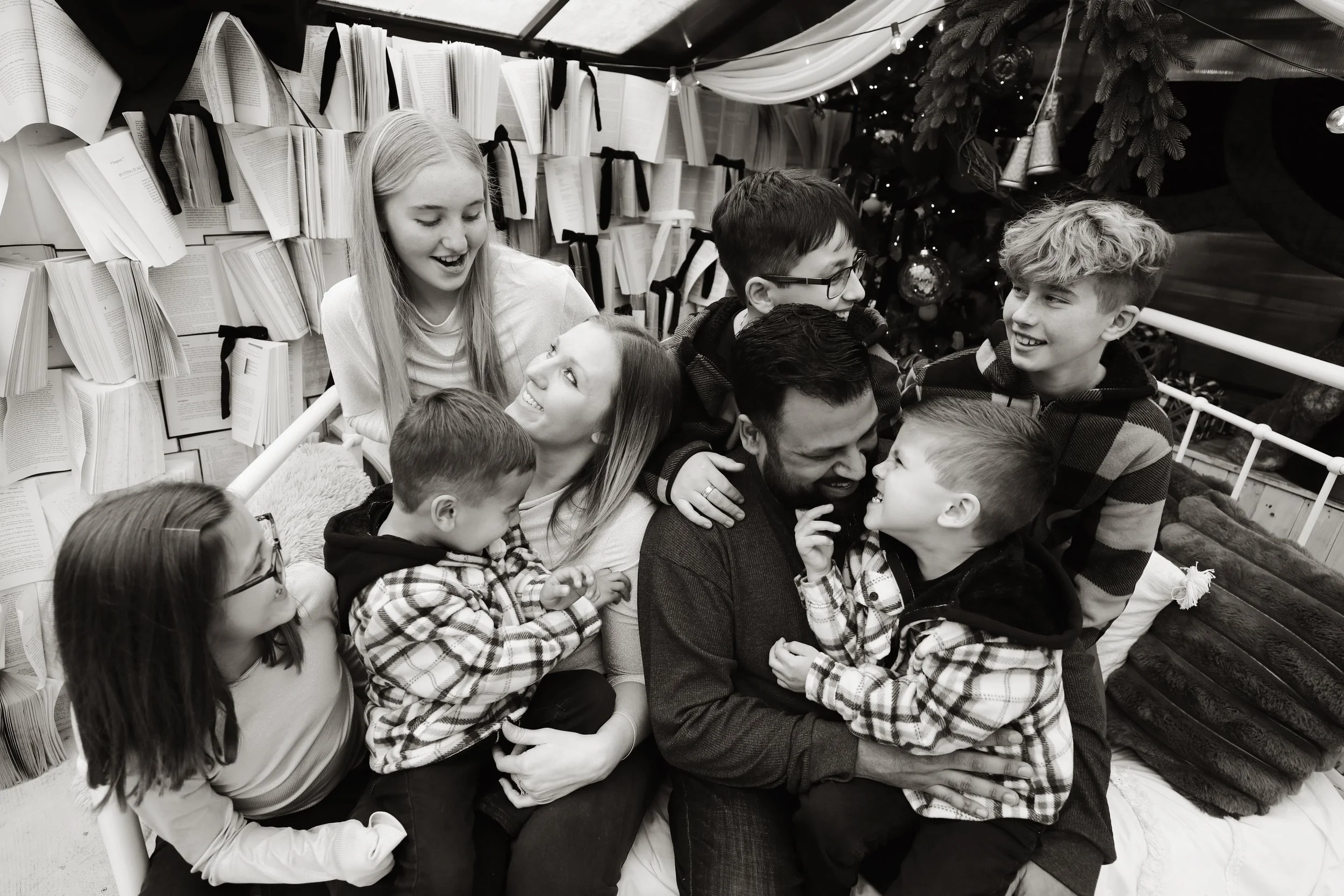 A group of children and an adult man smiling and playing together on a bed, surrounded by books and holiday decorations in a cozy room.