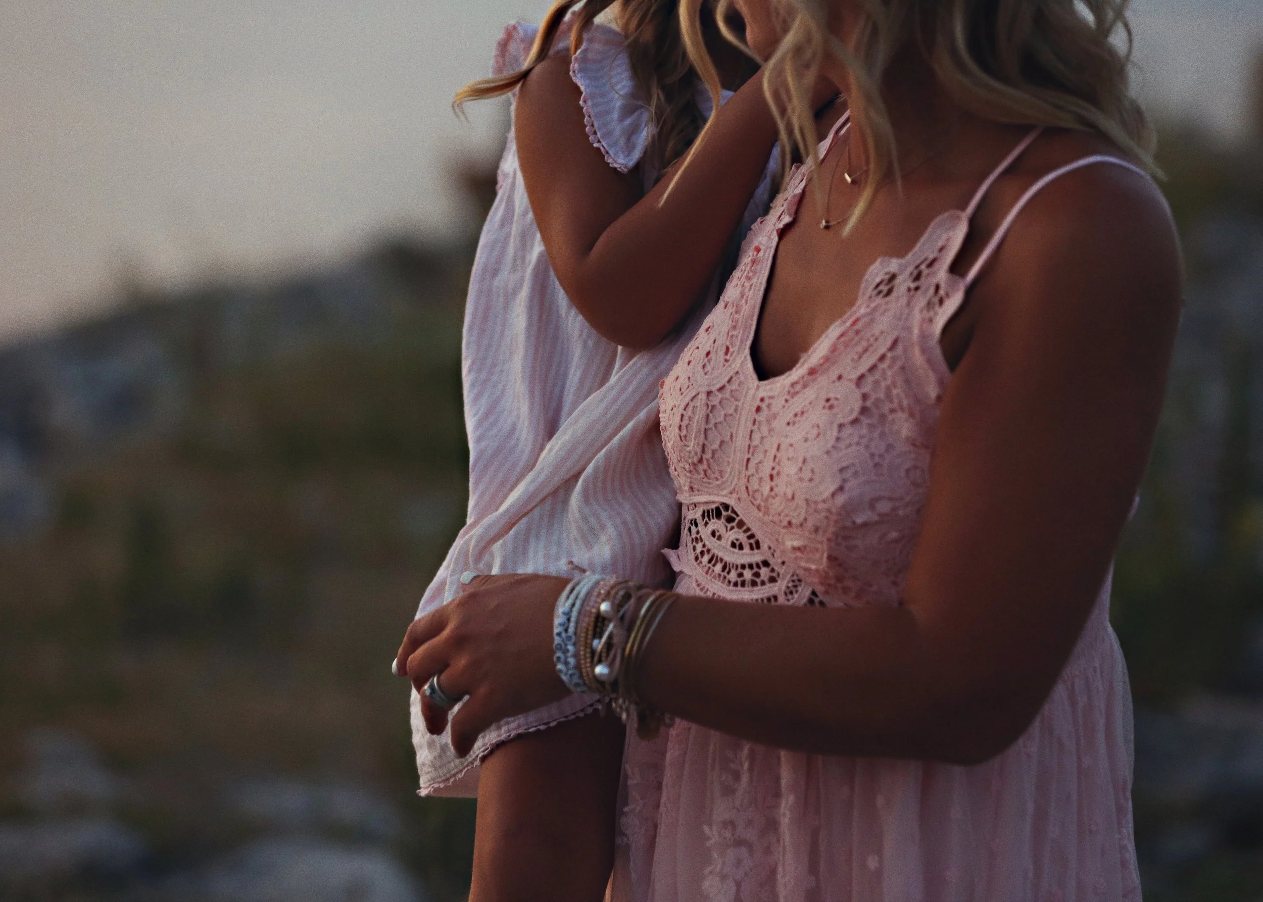 A woman in a pink lacy dress holding a child in a white dress on a beach at sunset.