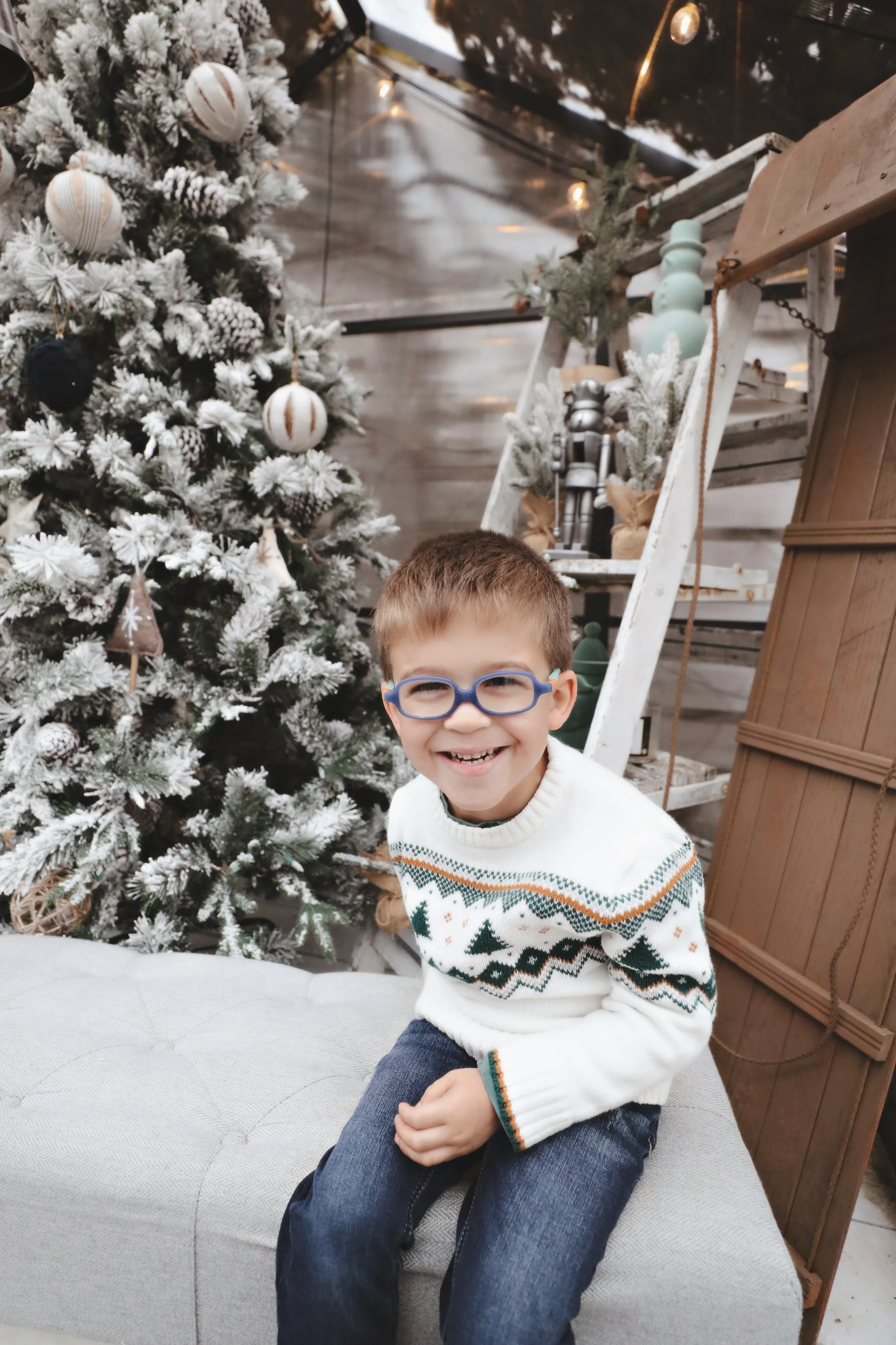 A young boy with glasses sitting on a gray ottoman in front of a decorated Christmas tree with white and metallic ornaments, smiling.