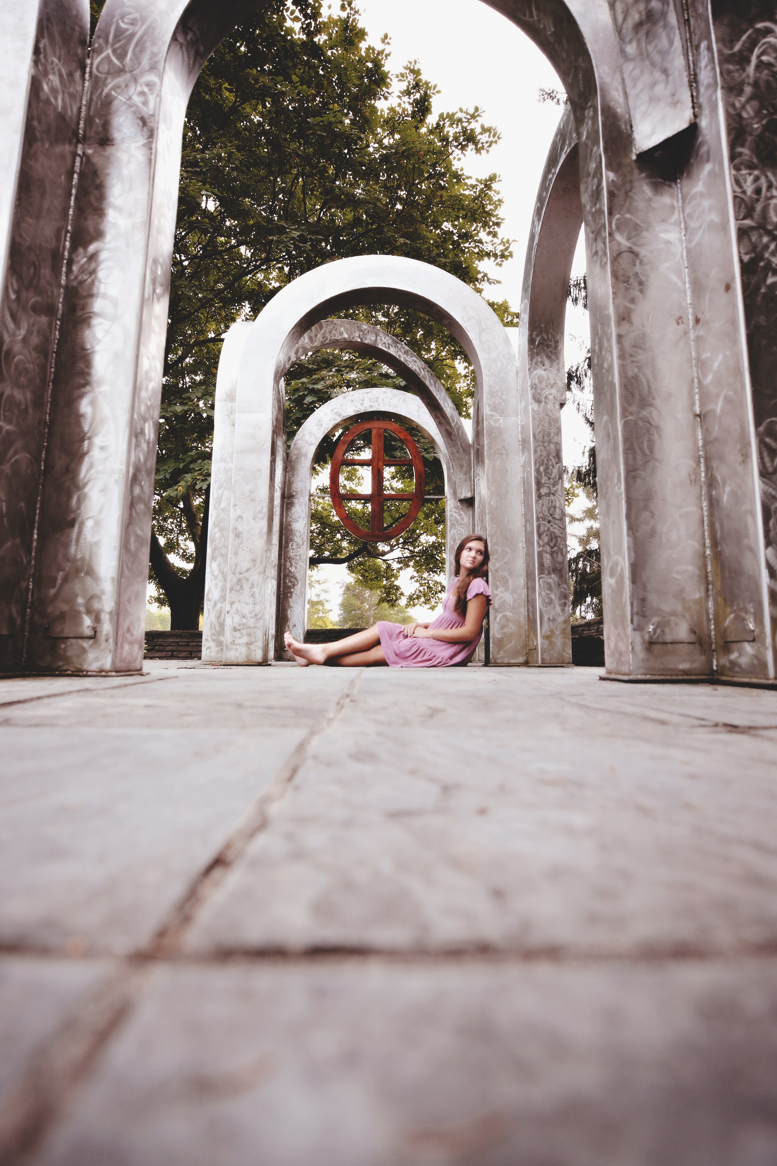 A young girl in a pink dress sitting on the ground underneath a series of arched metal structures in an outdoor setting with trees in the background.