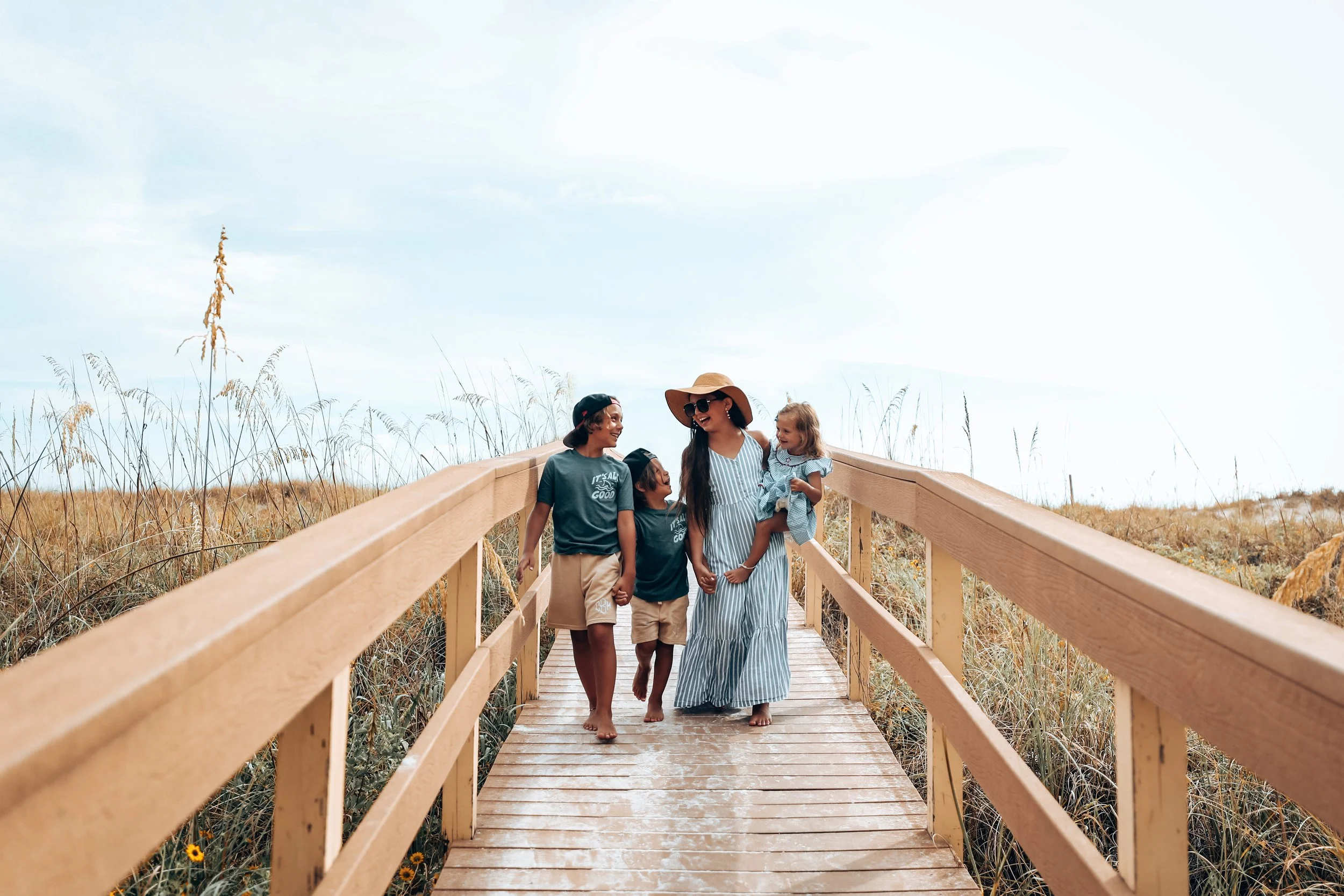 A woman in a blue and white striped dress and large sunhat holding a young girl, walking on a wooden boardwalk with three children in casual clothing, surrounded by tall grasses and a bright sky.