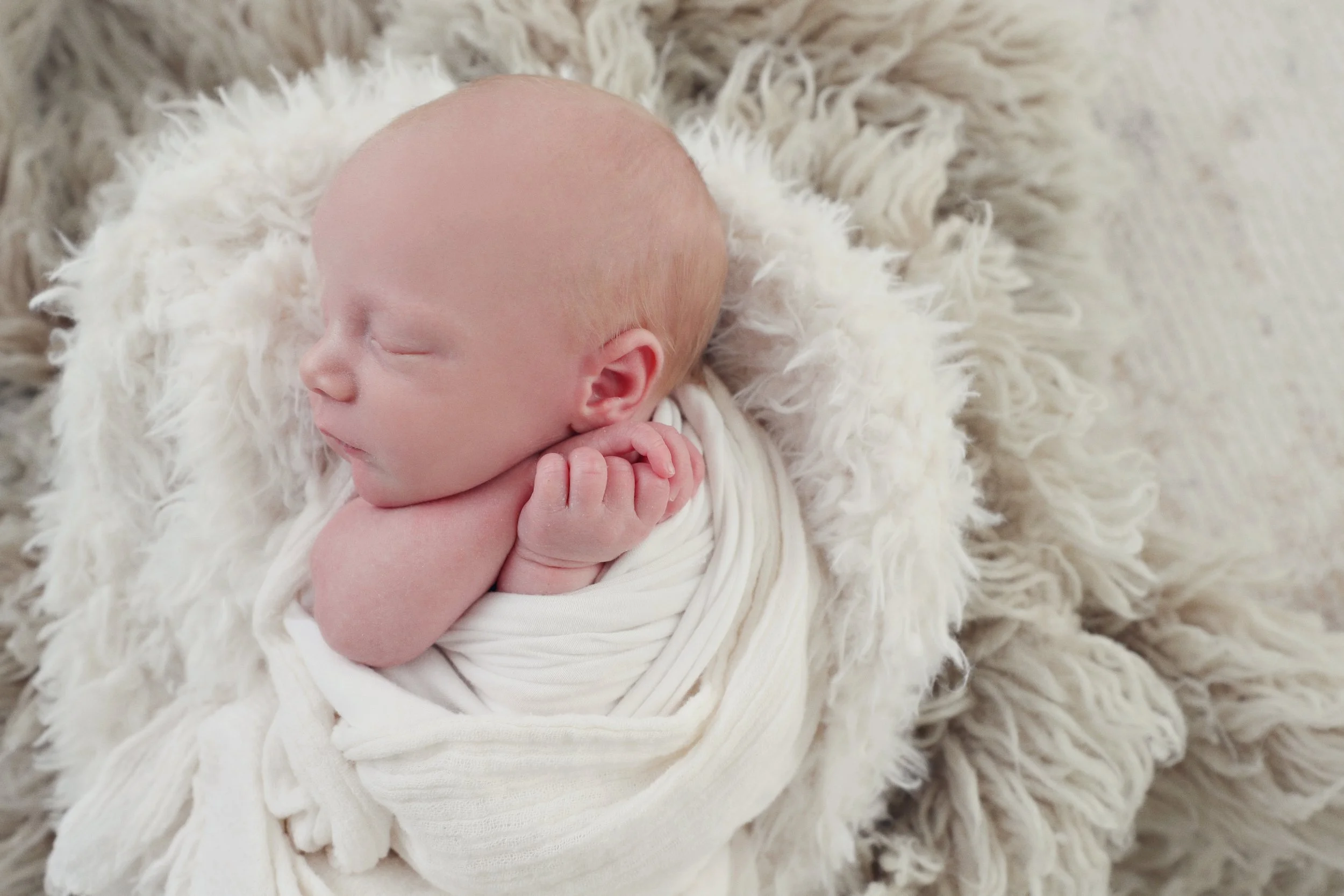 A sleeping newborn baby wrapped in a white blanket, resting on a fluffy cream-colored surface.