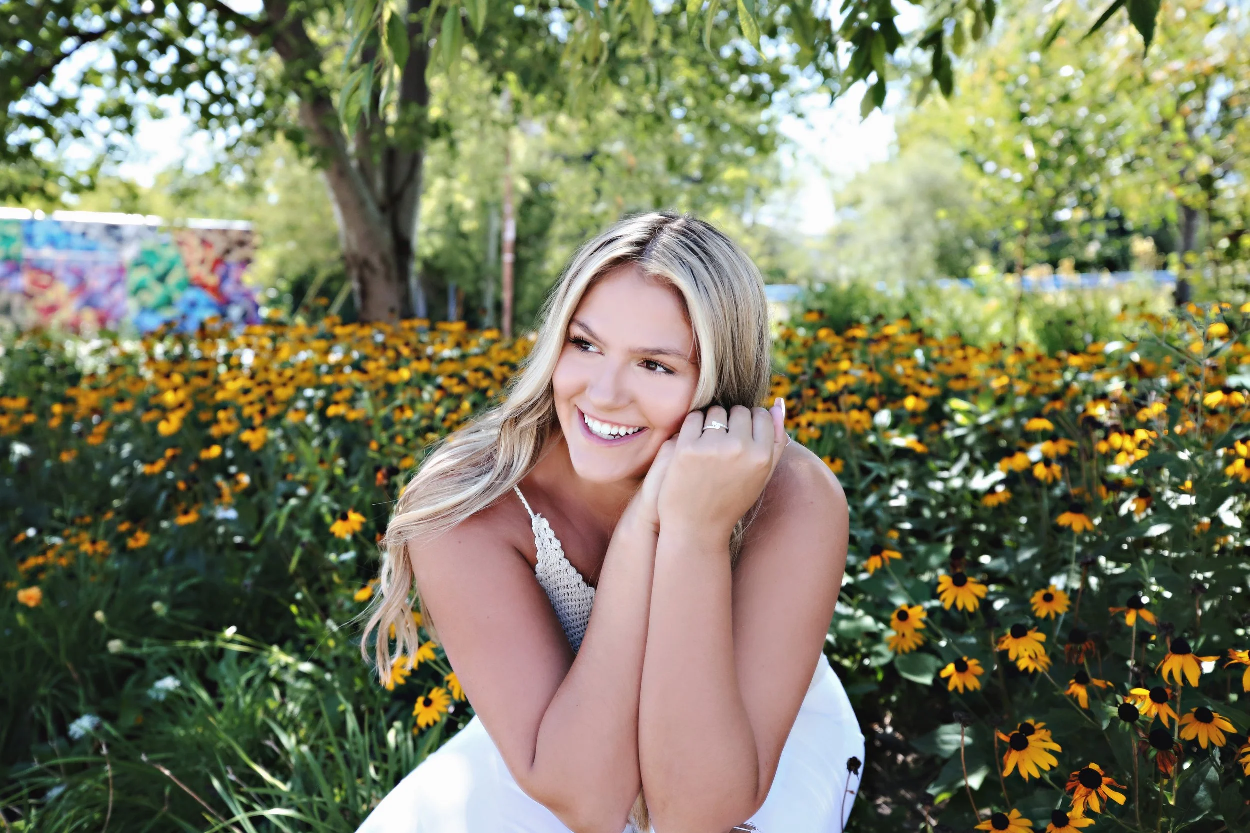 A woman with blonde hair smiling outdoors surrounded by yellow and orange flowers, trees, and greenery.