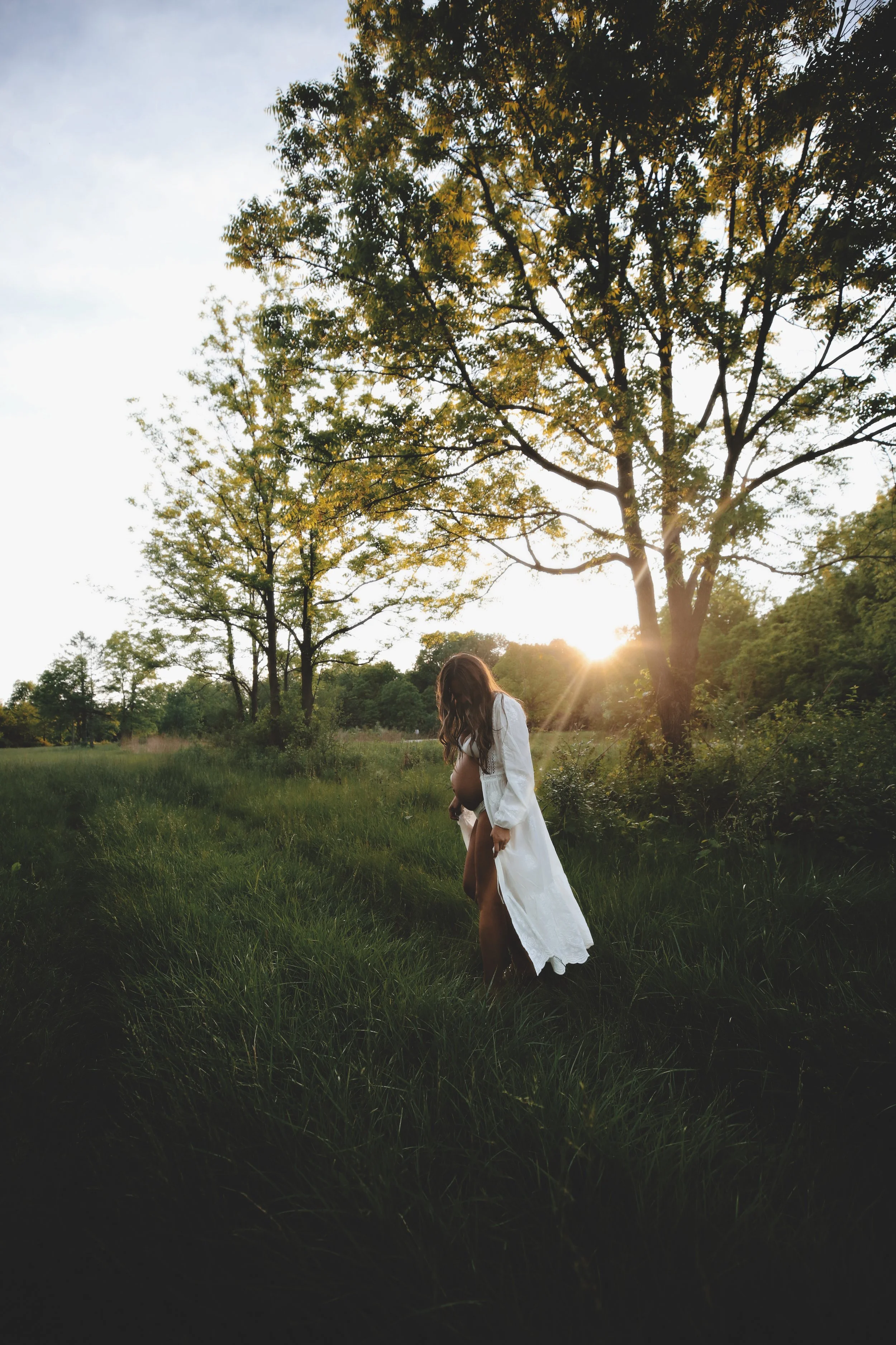 A pregnant woman in a white dress walking through a grassy field at sunset with trees in the background.