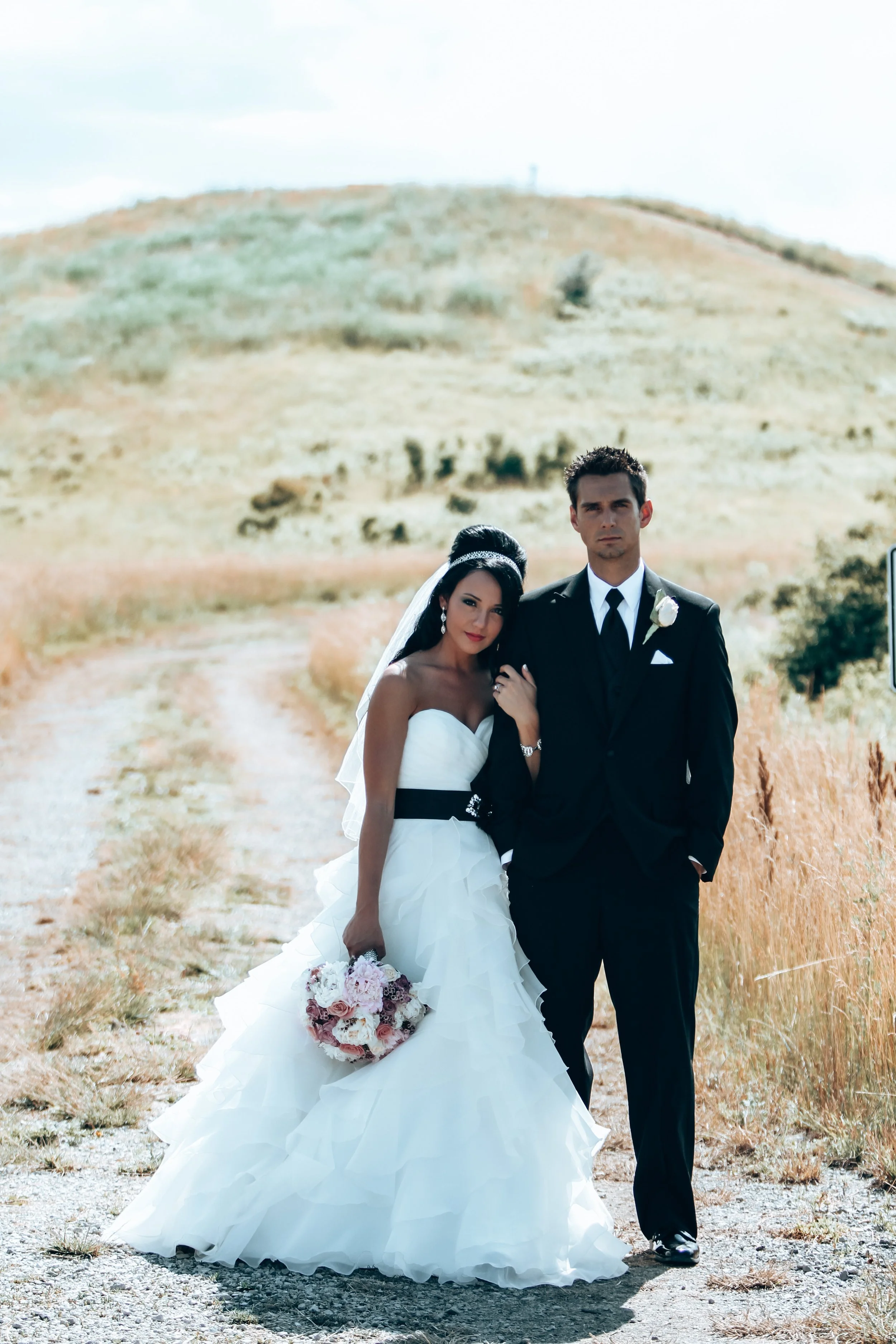 A bride and groom standing on a dirt path in a rural outdoor setting, with the bride holding a bouquet of flowers.