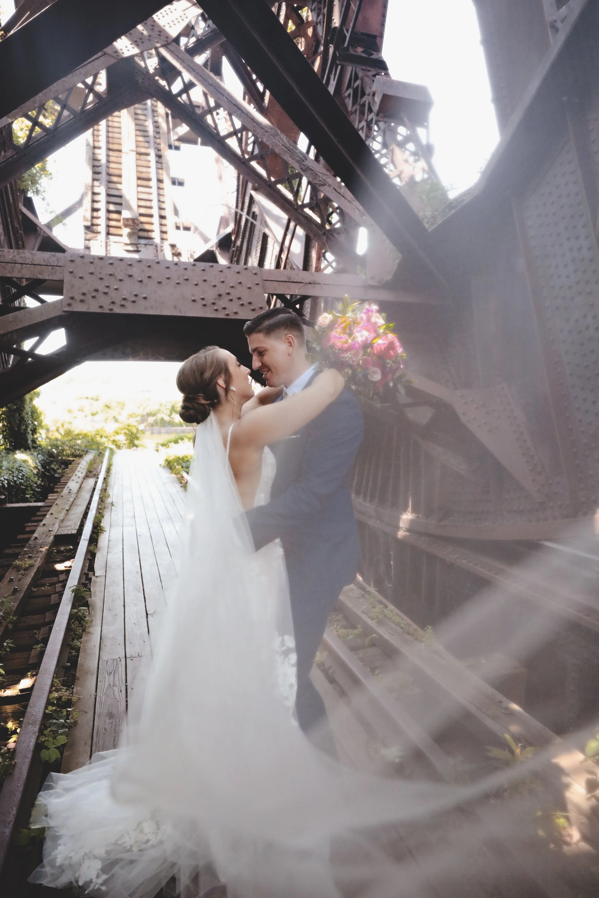 Bride and groom embracing under the Eiffel Tower structure, with the bride holding a bouquet, on a wooden pathway.