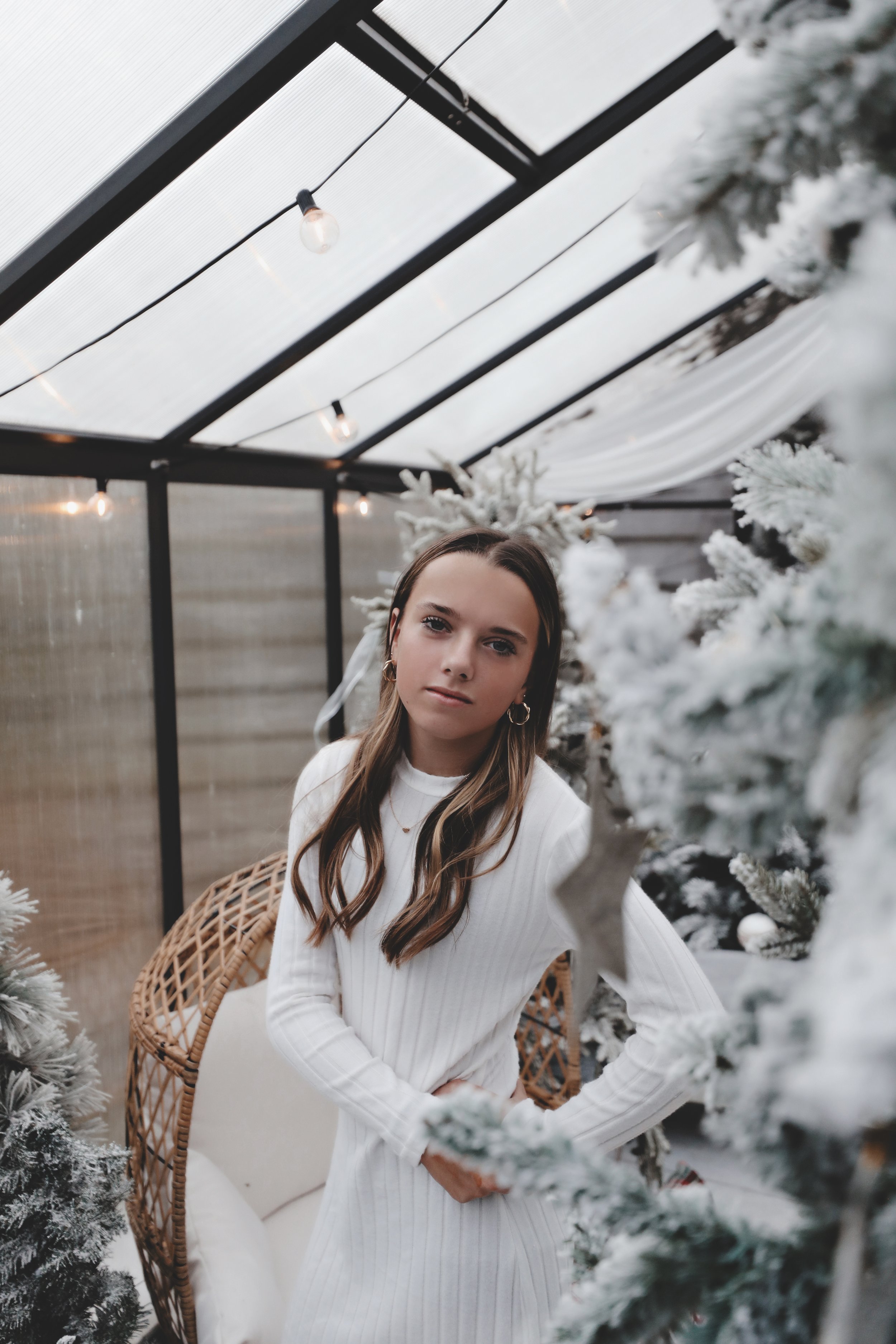 A young woman with long brown hair, wearing a white sweater dress, stands among snow-covered Christmas trees in a glass greenhouse decorated with string lights.