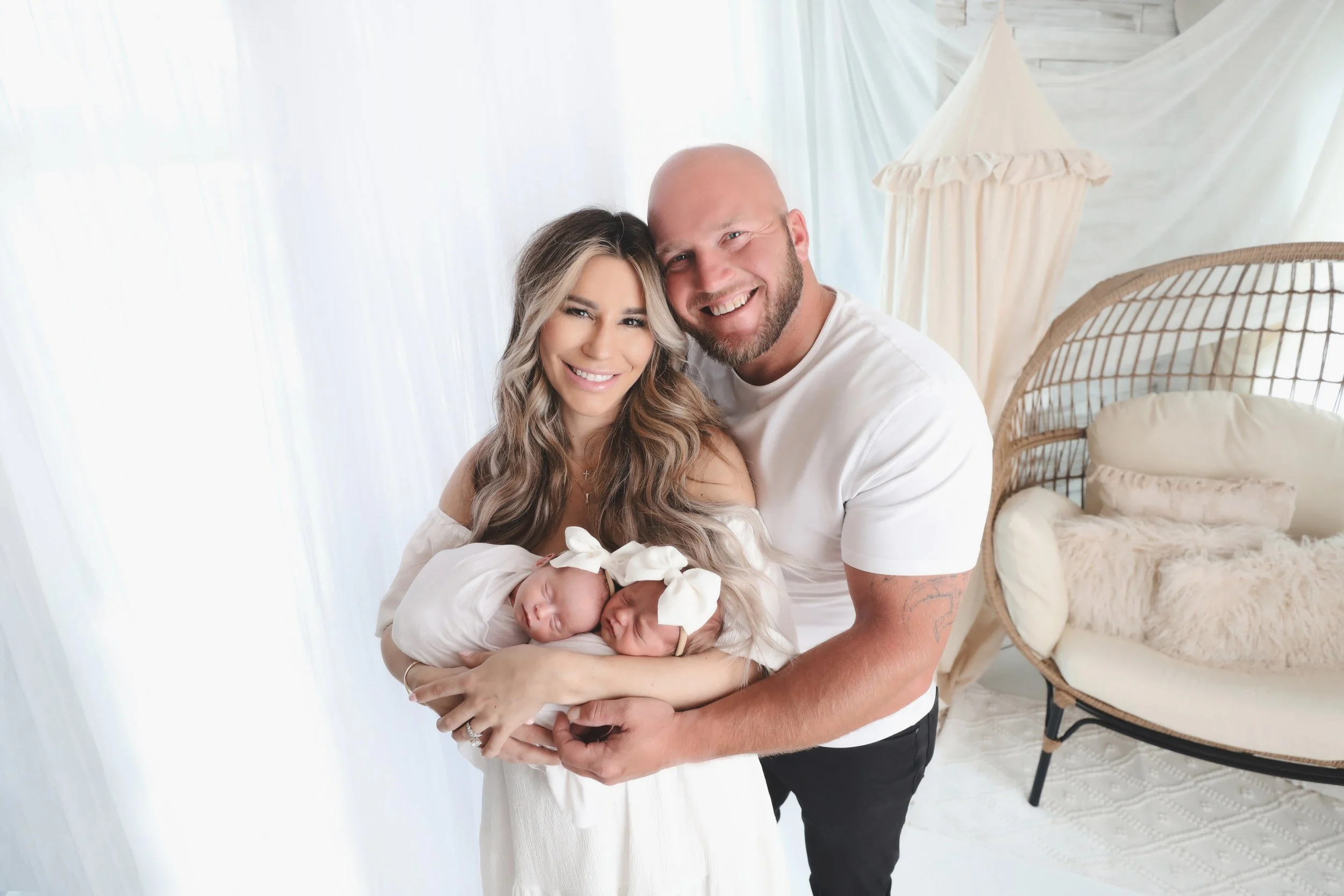A happy family holding two newborn twins in a bright, cozy room with white decor, including a cushioned chair and a canopy bed.