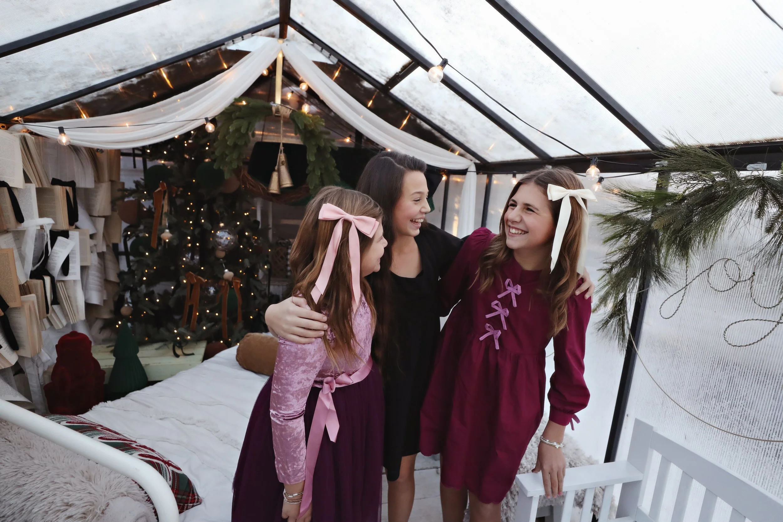 Three young girls with long hair, wearing dresses and bows, standing together inside a decorated glass greenhouse, smiling and embracing each other during Christmas.