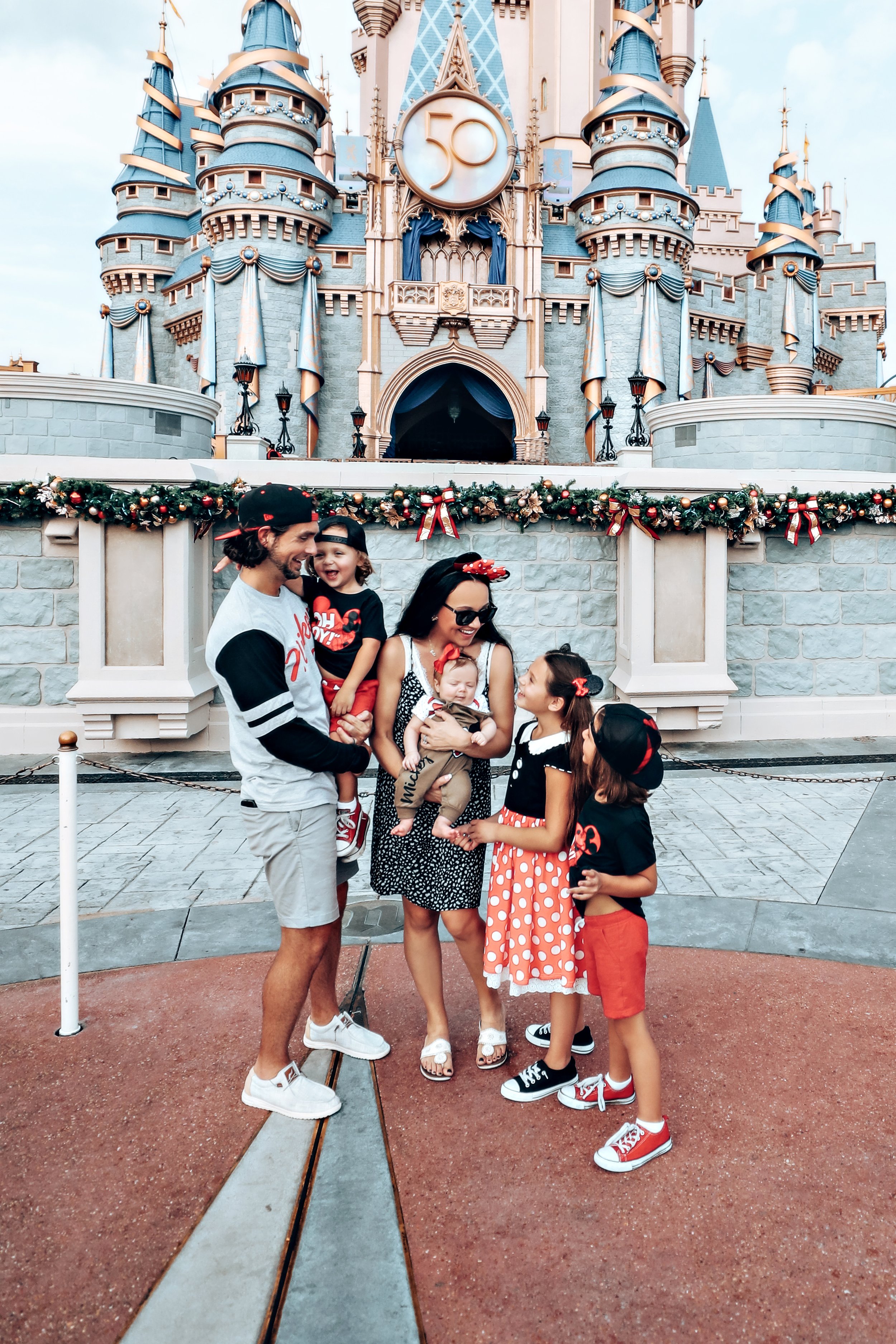 A family with three children celebrating in front of Sleeping Beauty's castle at Disneyland, decorated for Christmas with garland and ribbons, with a large 50th anniversary badge displayed above the castle.