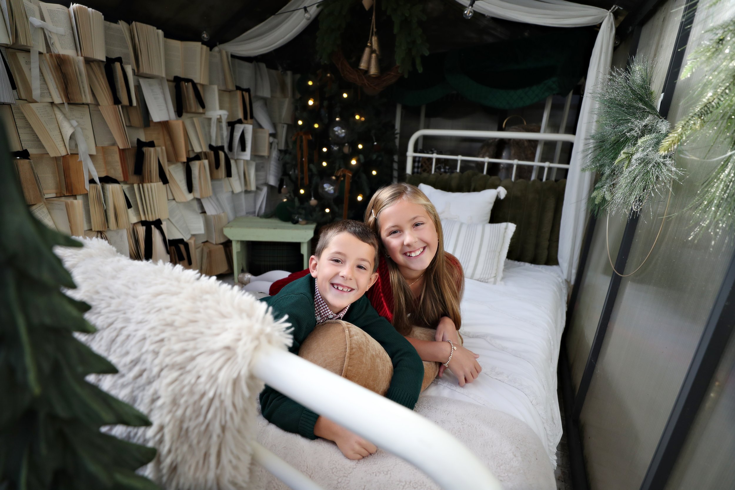 Two children, a boy and a girl, smiling and lying on a bed in a cozy, decorated room with bookshelves, a Christmas tree, and holiday decorations.