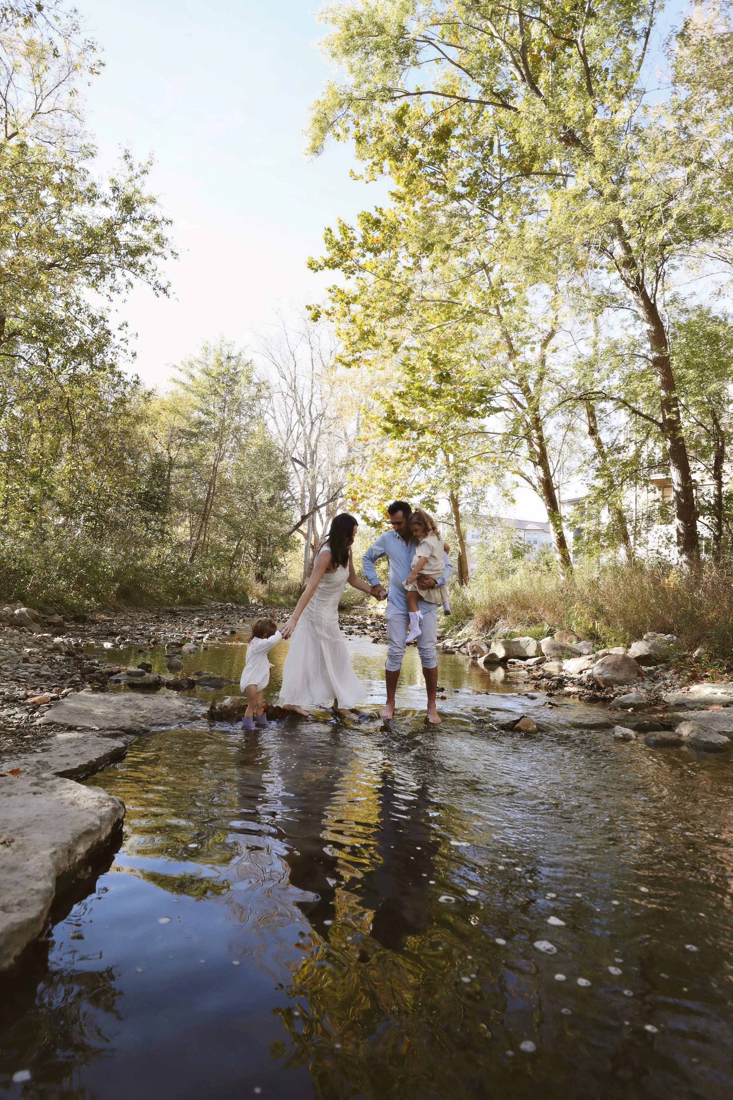 Family of four wading through a creek surrounded by trees, holding hands and enjoying a sunny day outdoors.