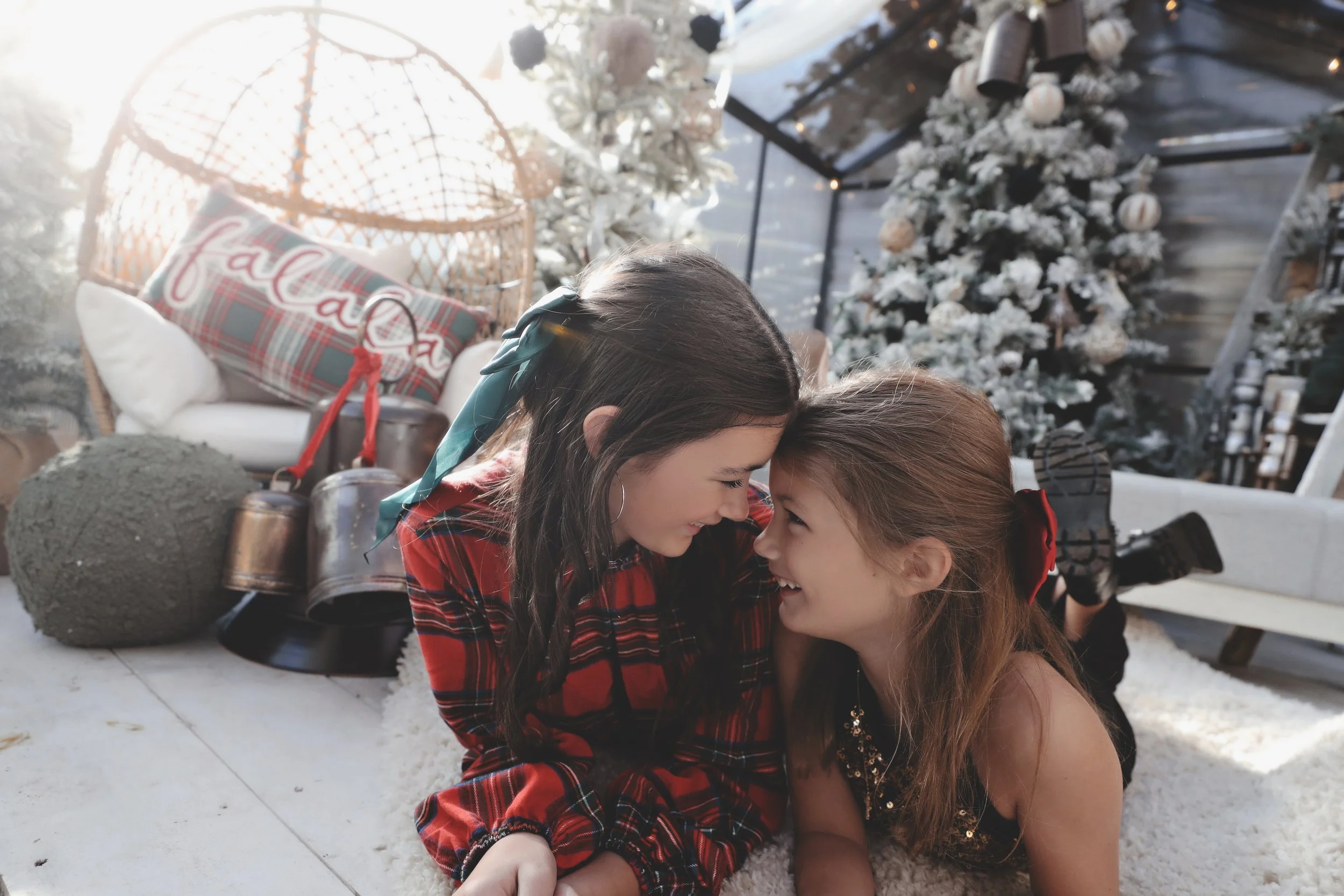 Two young girls smiling and touching foreheads in a festive Christmas setting with decorated trees, a wicker chair, and holiday decorations.