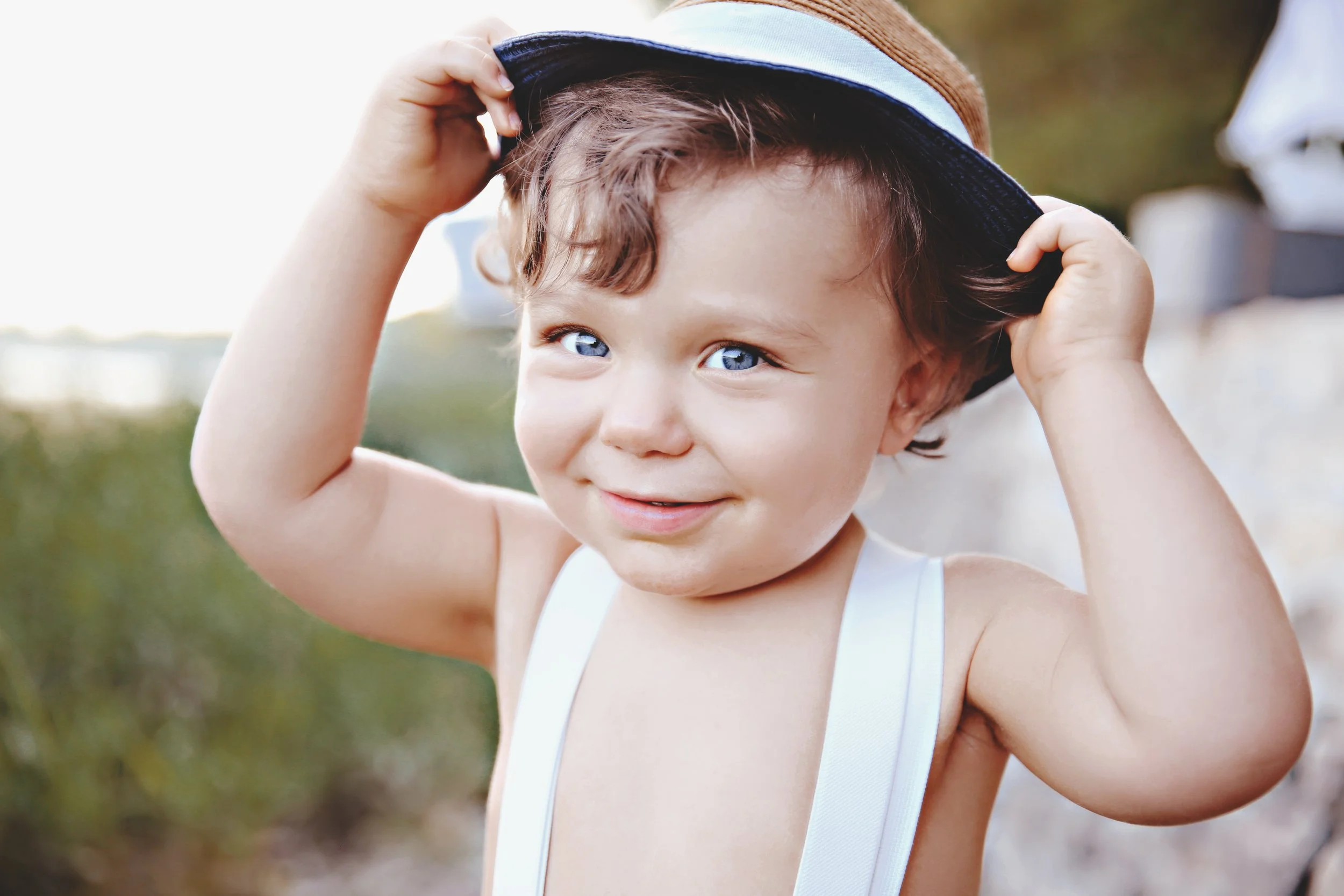 A young boy with curly brown hair and blue eyes adjusting a brown and blue wide-brimmed hat, wearing white suspenders, outdoors on a sunny day, smiling.