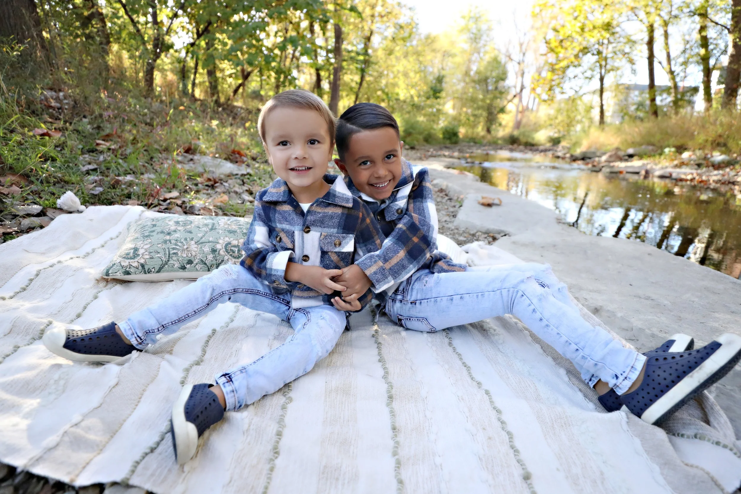 Two young boys sitting on a blanket by a river in a forest during autumn, smiling and holding hands.