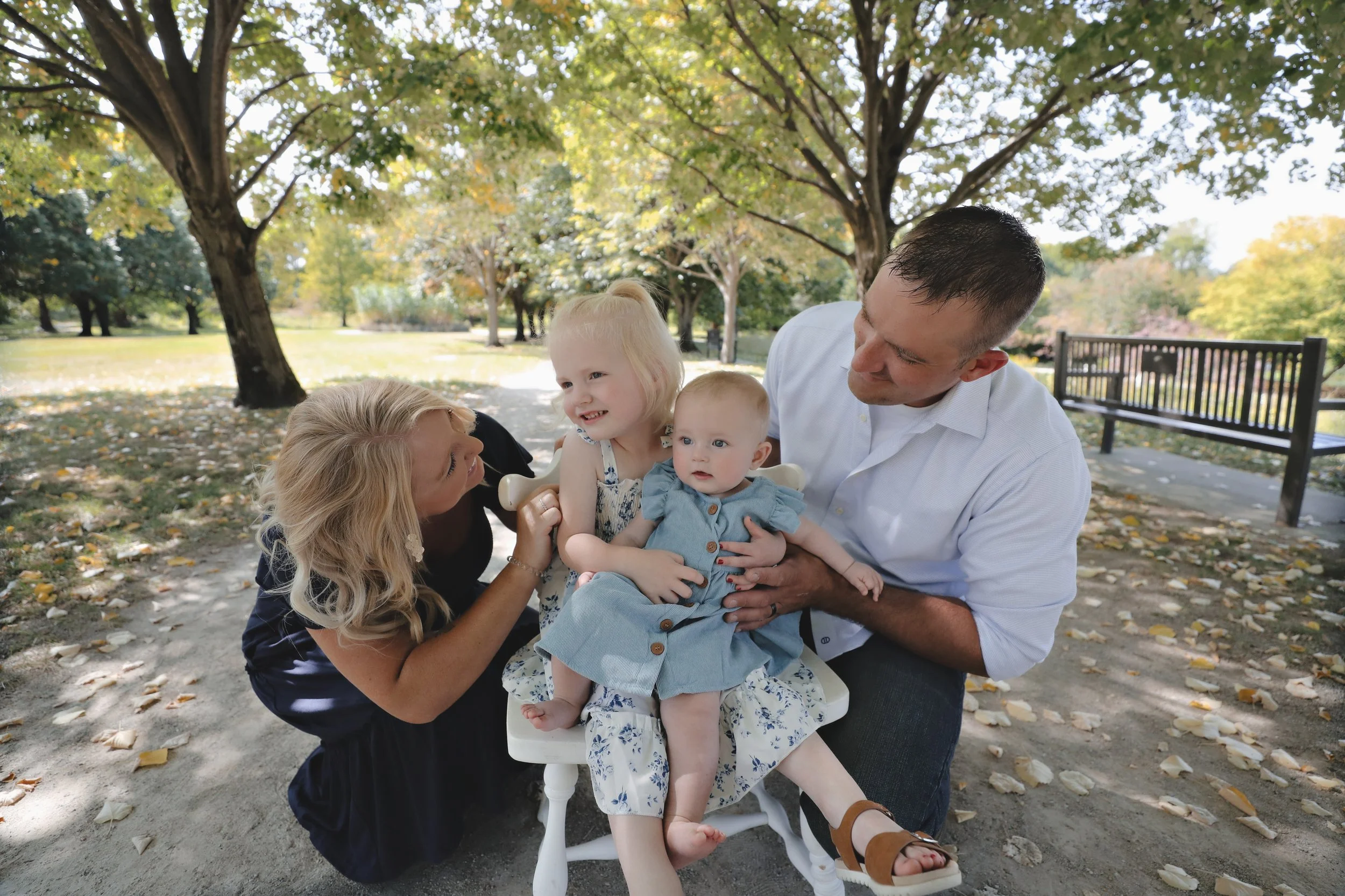 Family with a mother, father, and two young daughters sitting on a bench in a park with trees and fallen leaves during autumn.