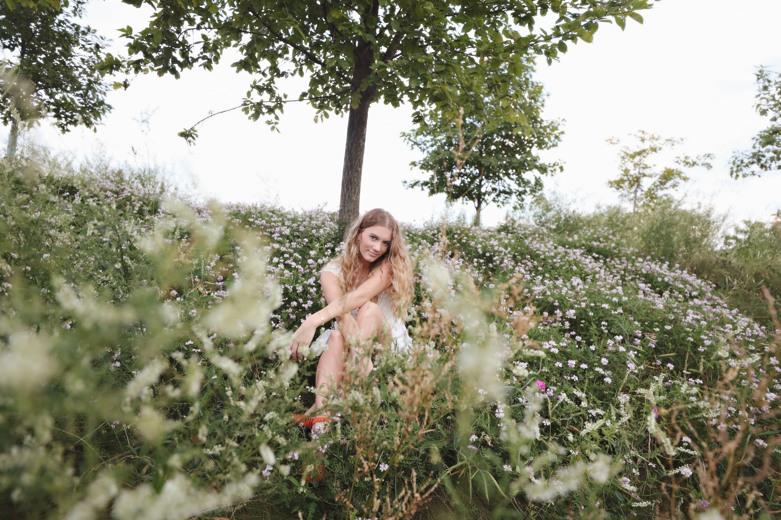 A young woman with long curly blonde hair sitting among flowering bushes with white and pink flowers, outdoors on a partly cloudy day, with trees and a green landscape in the background.