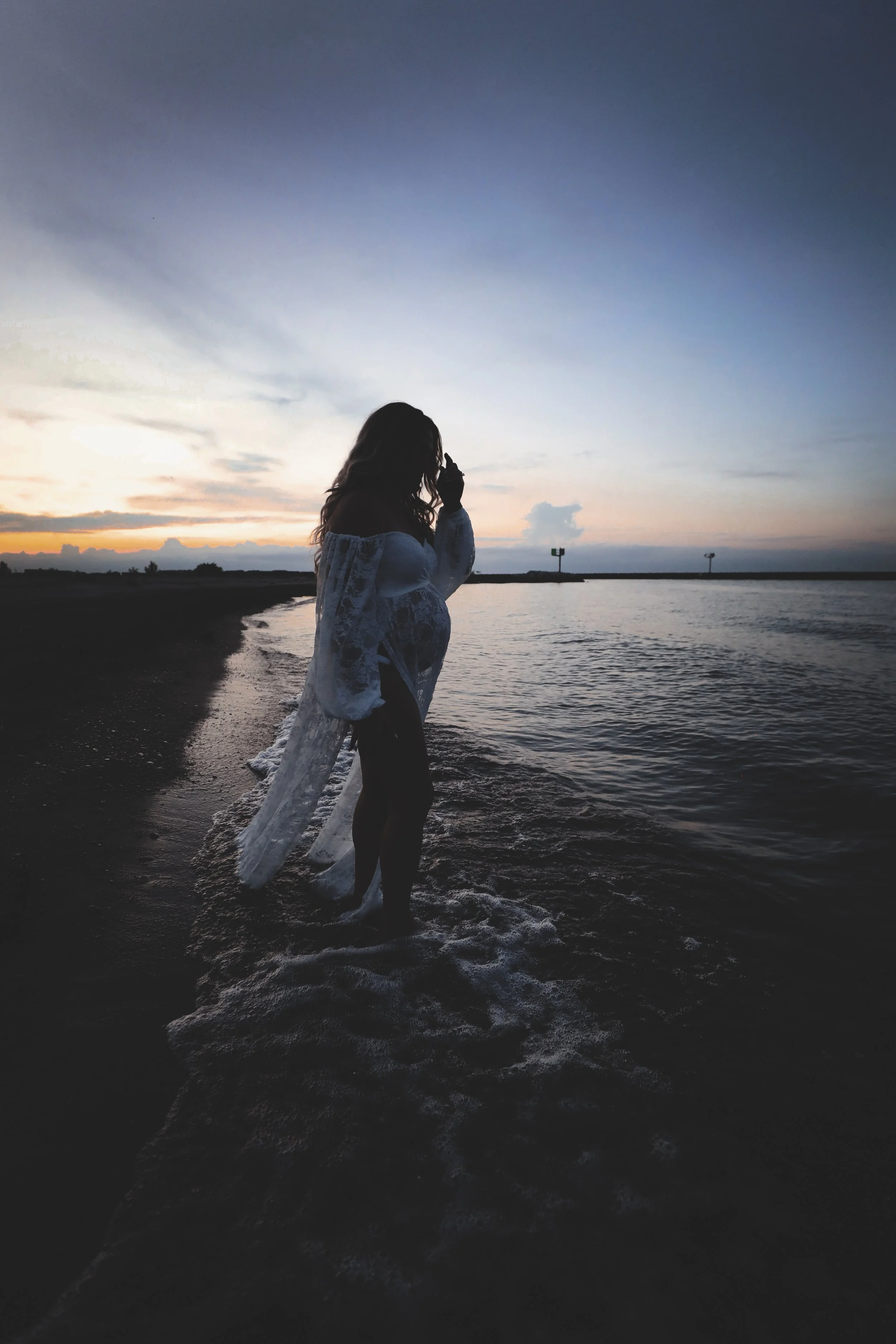 Silhouette of a pregnant woman standing in the shallow water at the beach during sunset with the sky showing orange, pink, and blue hues.