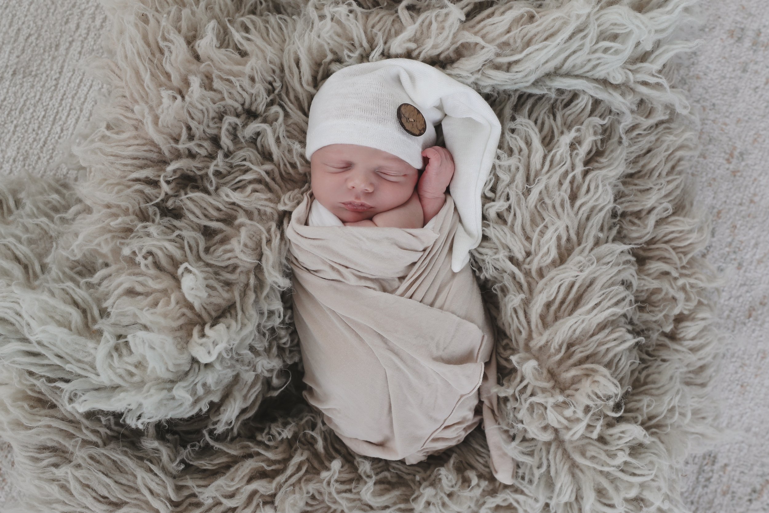 A newborn baby sleeping peacefully, swaddled in a beige blanket, wearing a white hat with a brown button, lying on a fluffy beige rug.