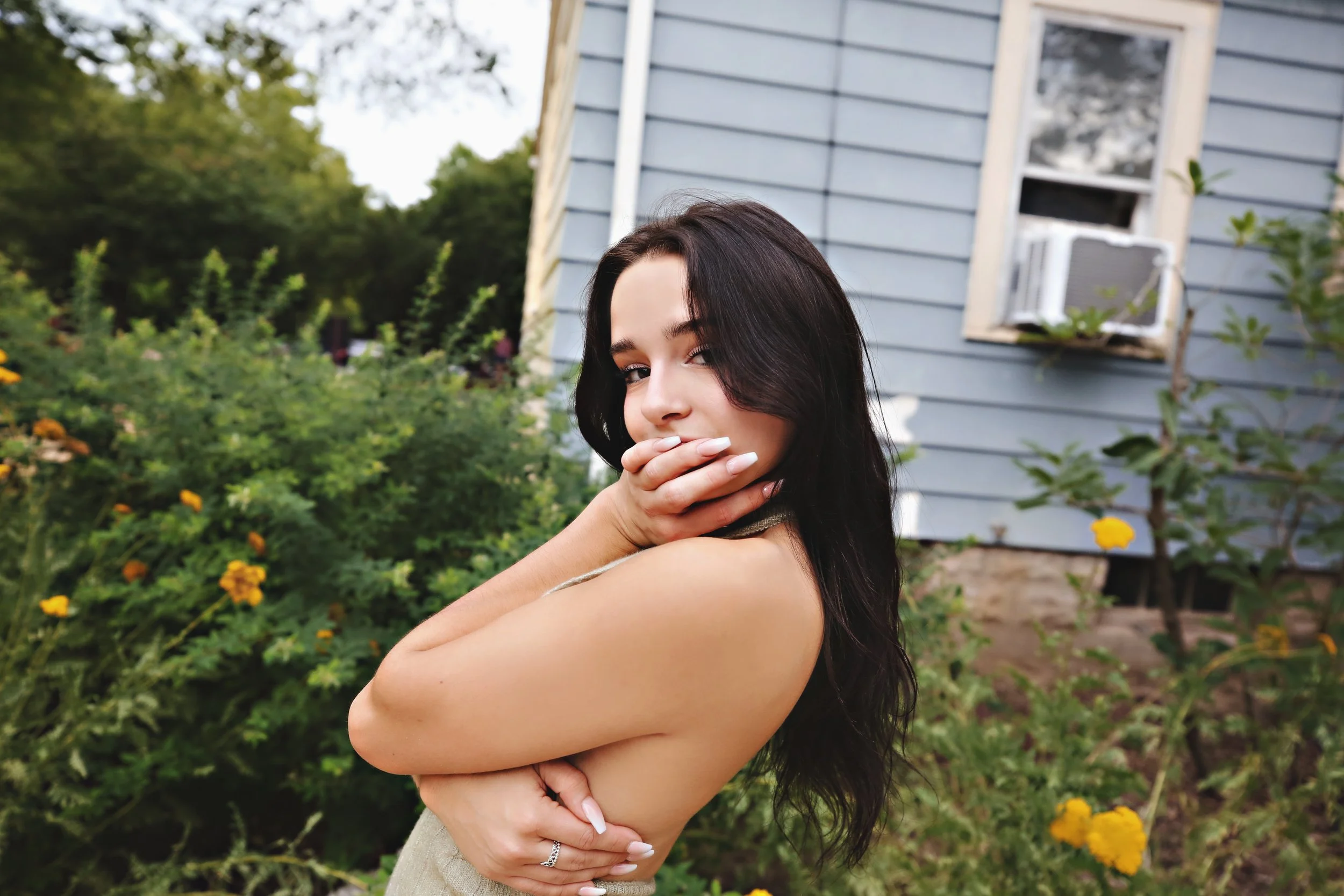 A woman with dark hair embracing herself in a garden with yellow flowers, with a light blue house and an off-white window air conditioning unit in the background.