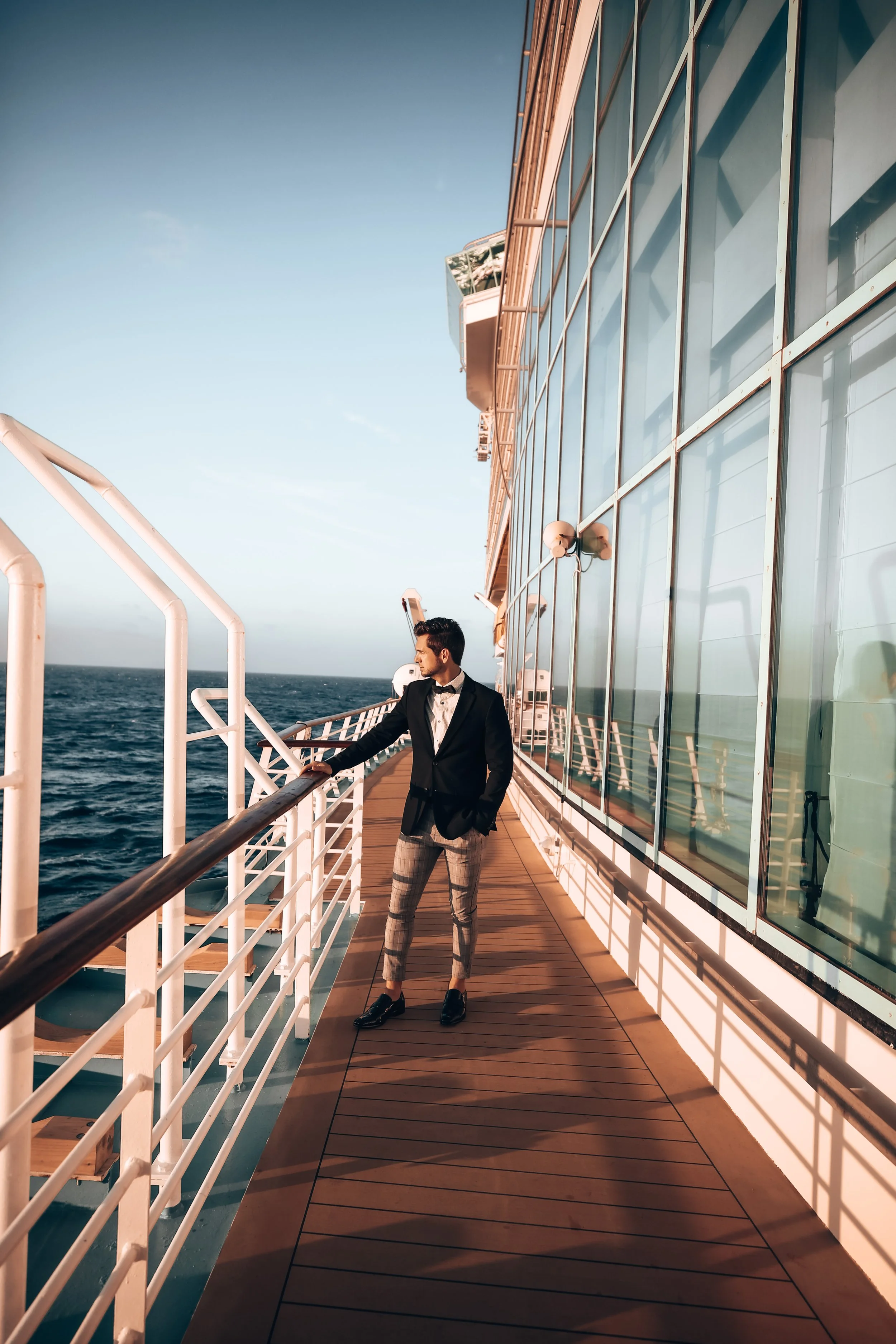 A man in a tuxedo with plaid pants standing on the deck of a ship, looking out at the ocean.