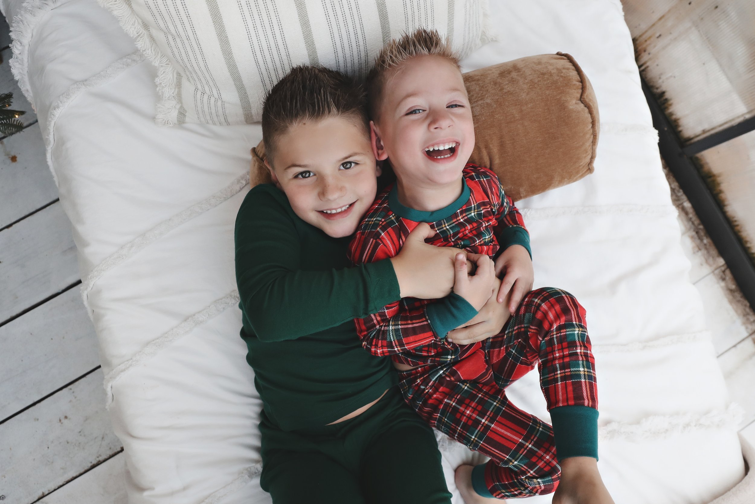 Two young boys smiling and laughing while lying on a bed, one in green pajamas and the other in red plaid pajamas, hugging each other.
