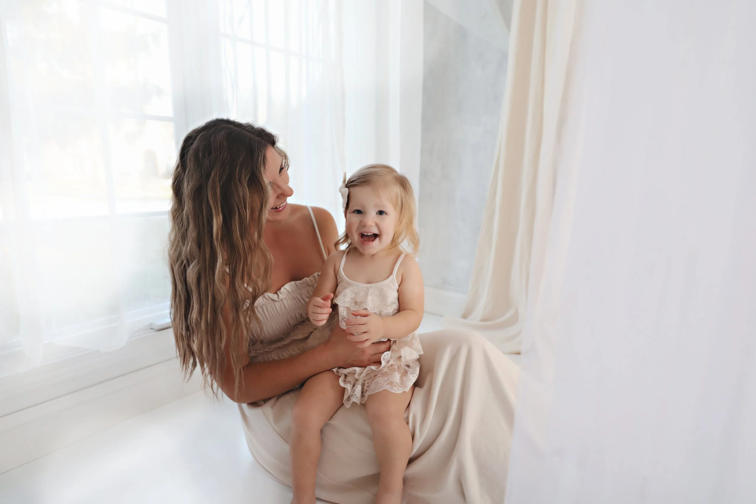 A woman and a young girl sitting by a large window with white curtains, smiling and laughing together.