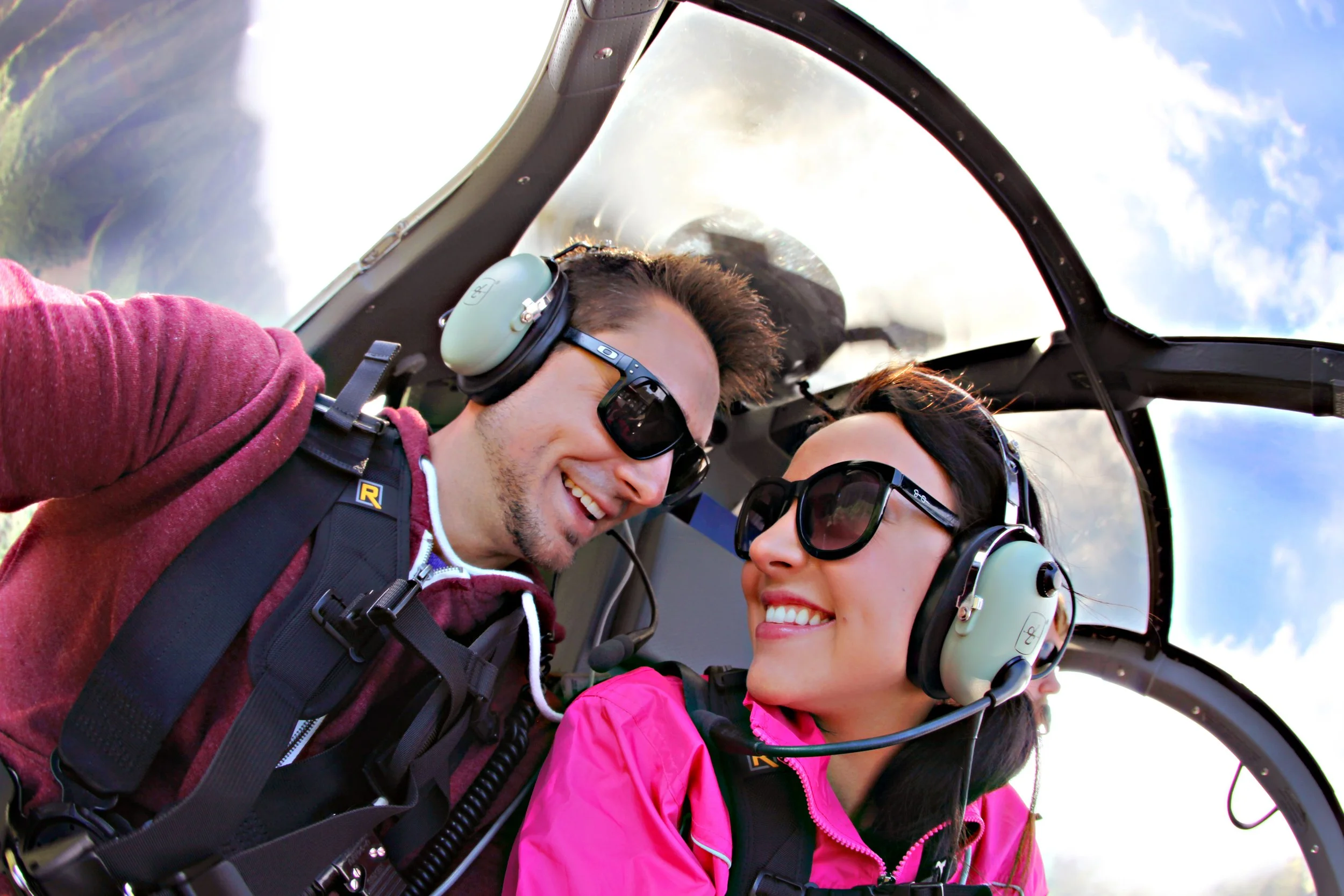 Two smiling pilots in a helicopter cockpit, both wearing sunglasses and headsets, with a blue sky and clouds visible behind them.