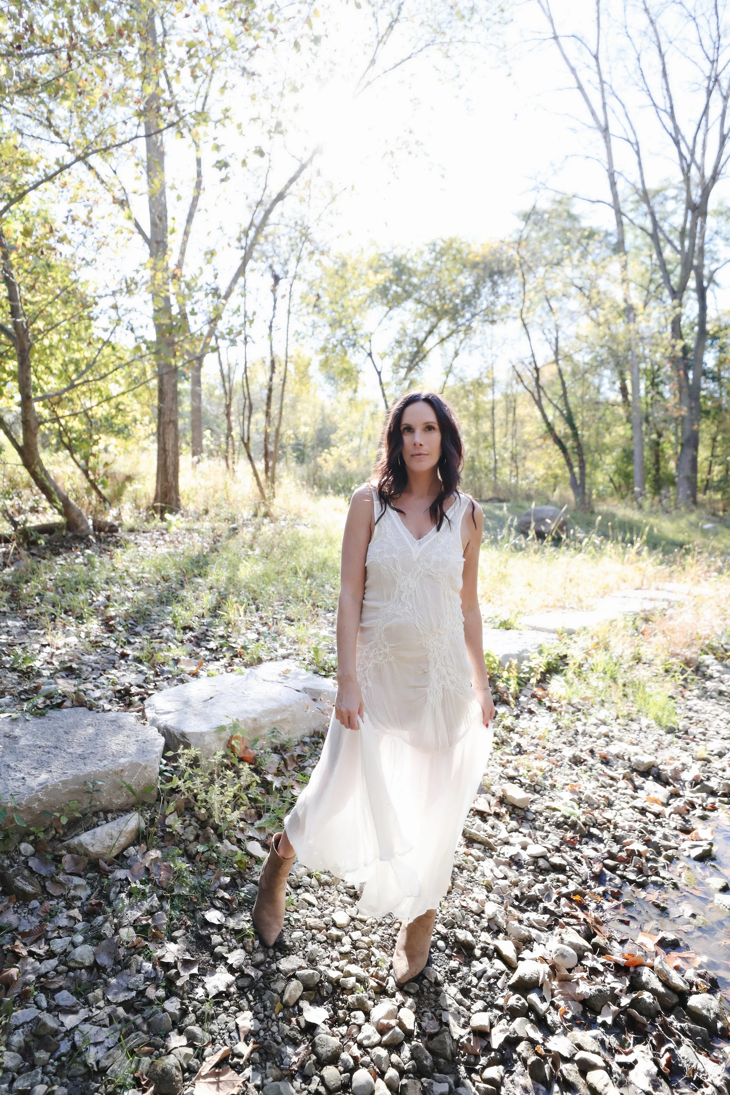 A woman in a long white dress and brown boots walking on a rocky path in a wooded area during daylight.