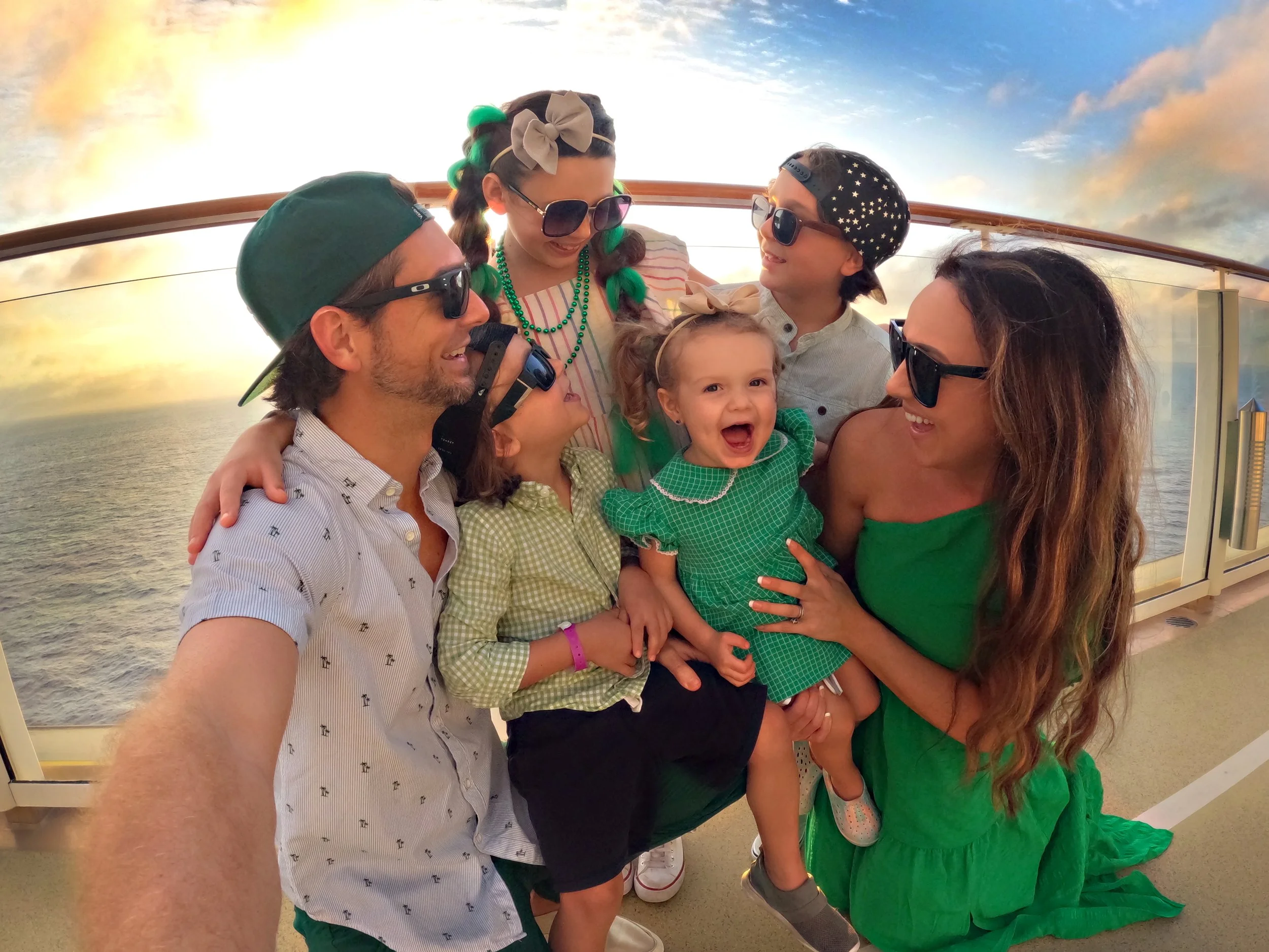 Family taking a group selfie on a cruise ship deck with ocean and sunset in the background, all wearing sunglasses and casual summer clothing.