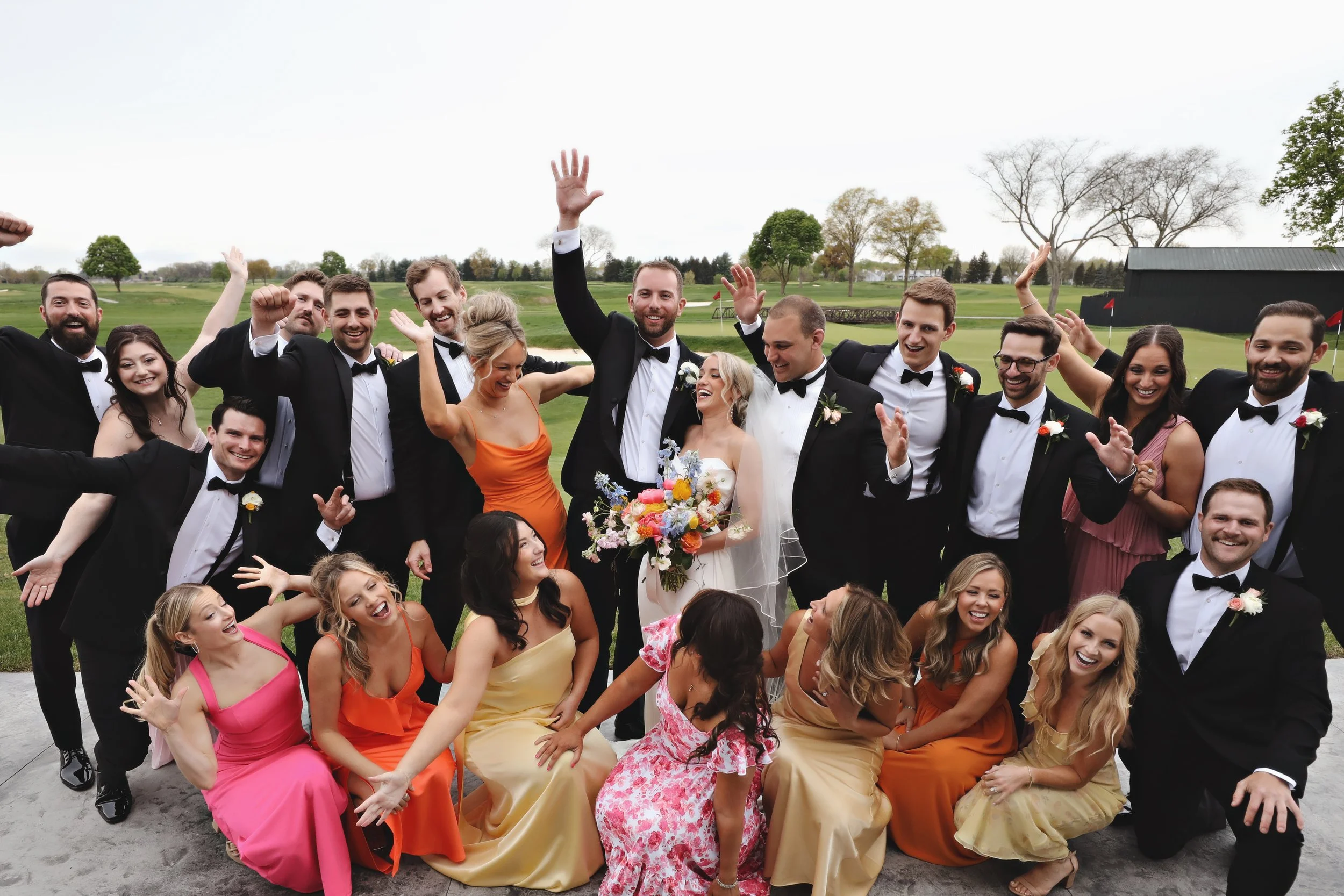 A group of people, including bridesmaids and groomsmen, celebrating outdoors on a golf course with trees and overcast sky in the background, at a wedding party.