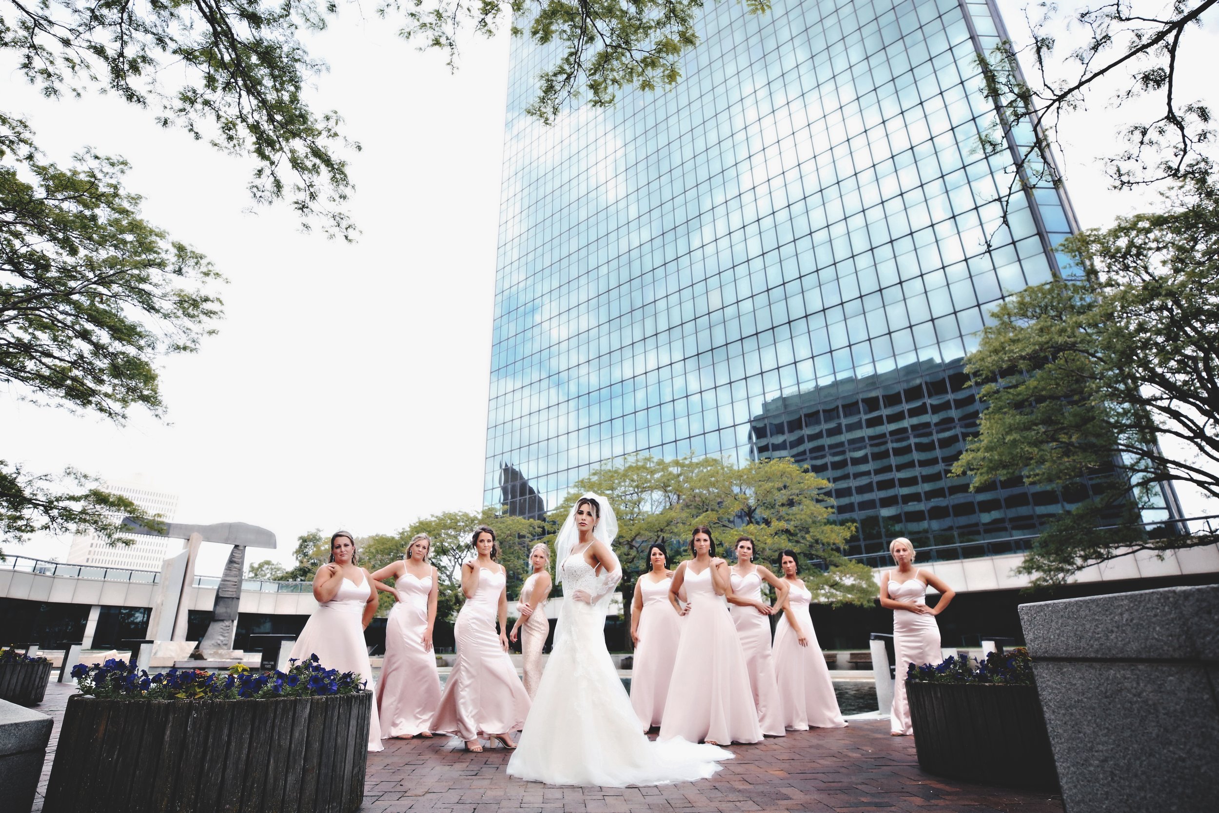 A bride and her bridesmaids are standing outdoors in front of a modern glass office building. The bride is in the center wearing a white wedding gown and veil, while the bridesmaids are in matching light pink dresses.