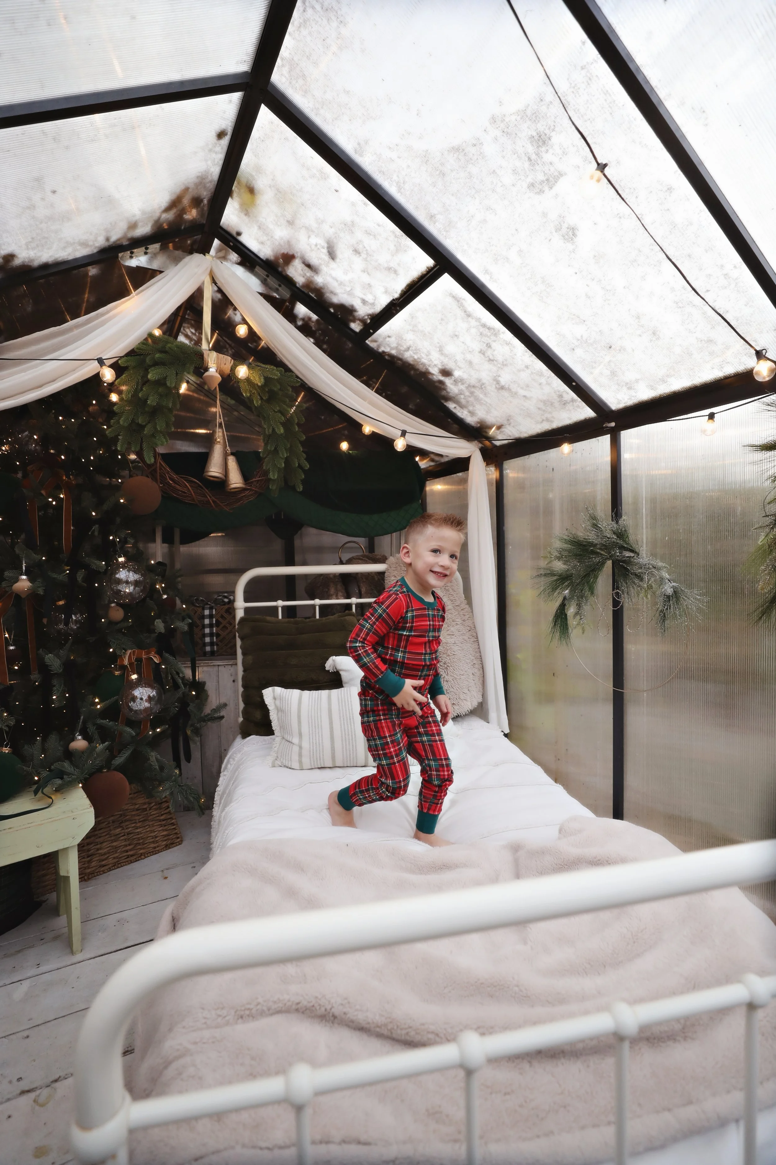 A young boy in red plaid pajamas jumping on a bed in a cozy, decorated room with holiday decorations and a Christmas tree.