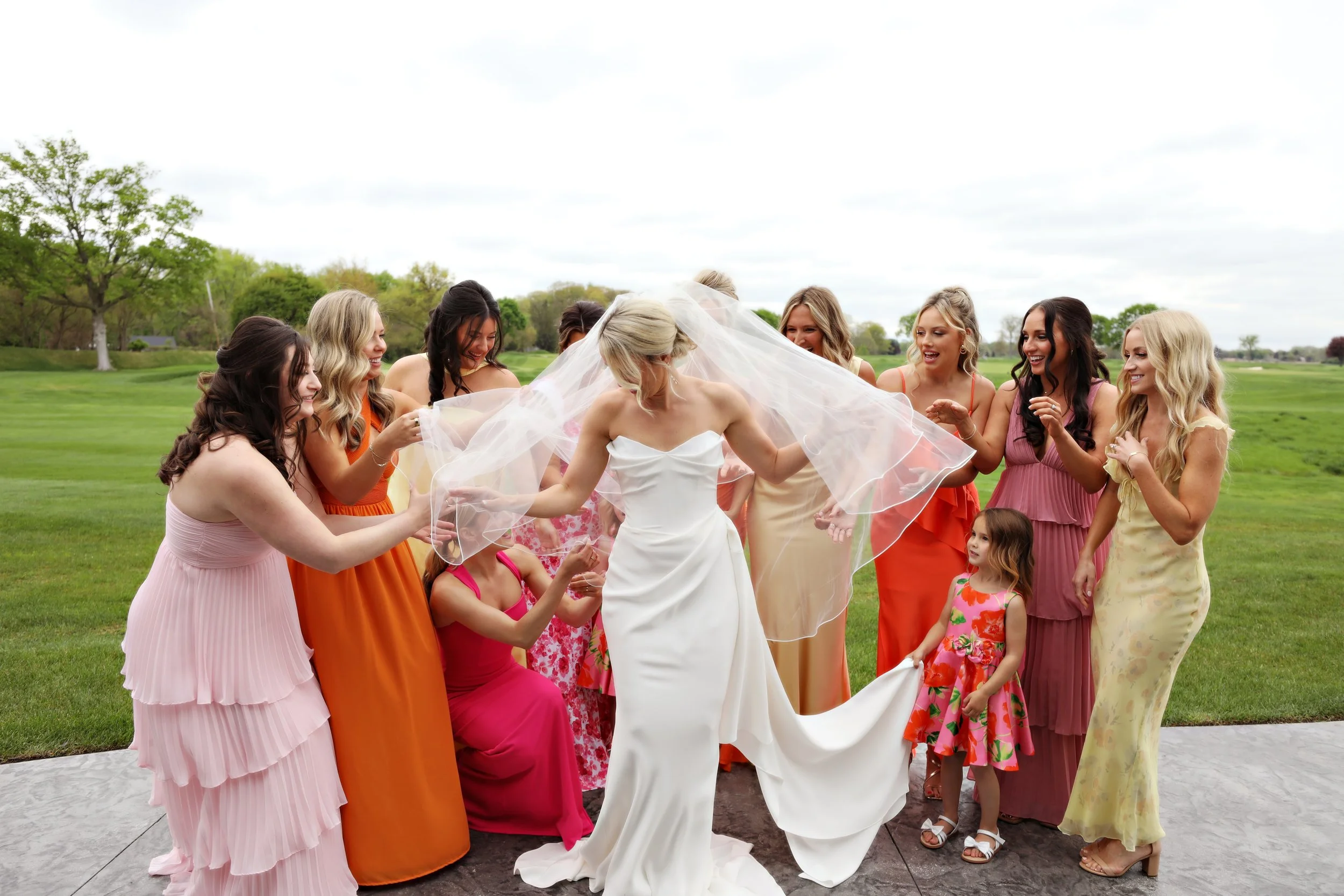 A bride in a white wedding gown holding a veil is surrounded by friends and flower girls on a grassy outdoor area on a cloudy day.