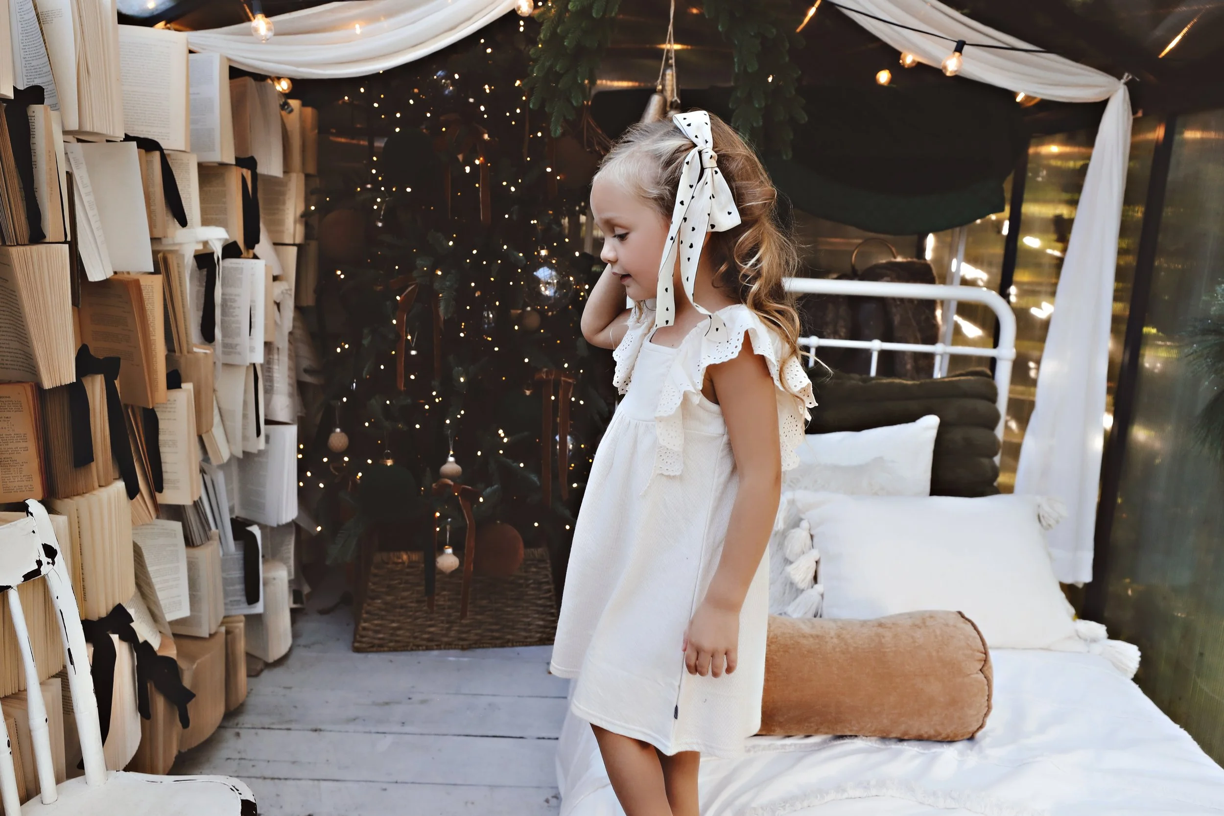 A young girl in a white dress with ruffled sleeves and a polka dot bow in her hair, standing indoors in front of a decorated Christmas tree with lights, near a bed with white pillows and a brown bolster, next to a book shelf filled with books.