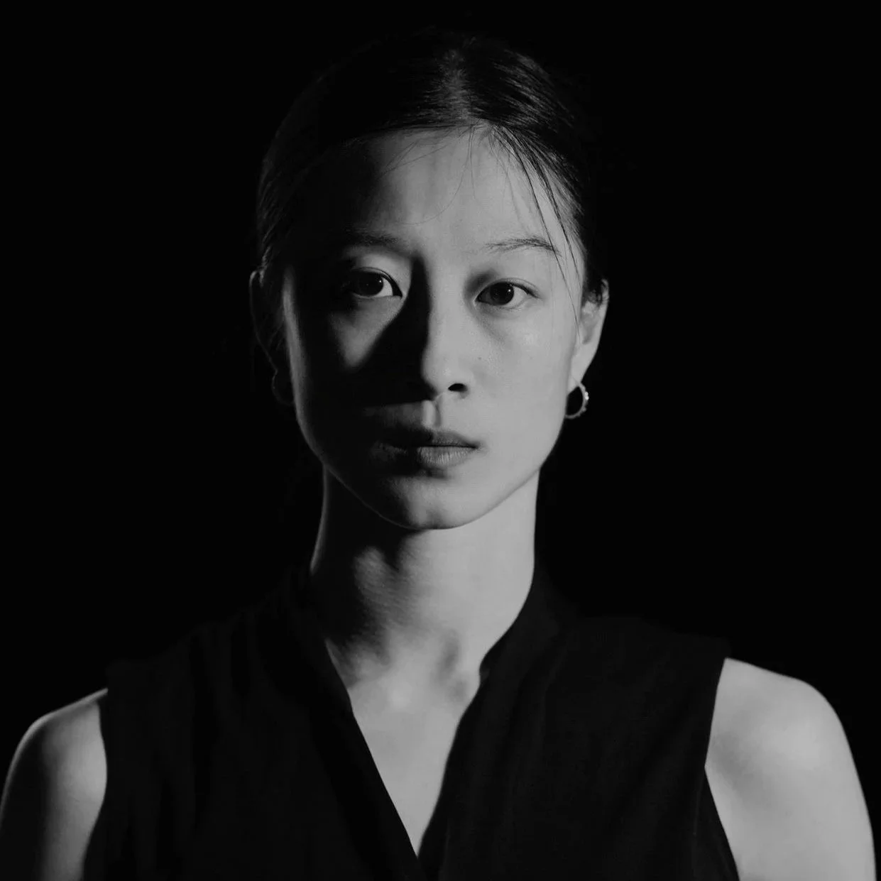 Schwarz-weiß Portrait einer jungen Frau mit kurzen Haaren, wearing einer schwarzen ärmellosen Bluse, gegen schwarzen Hintergrund.
