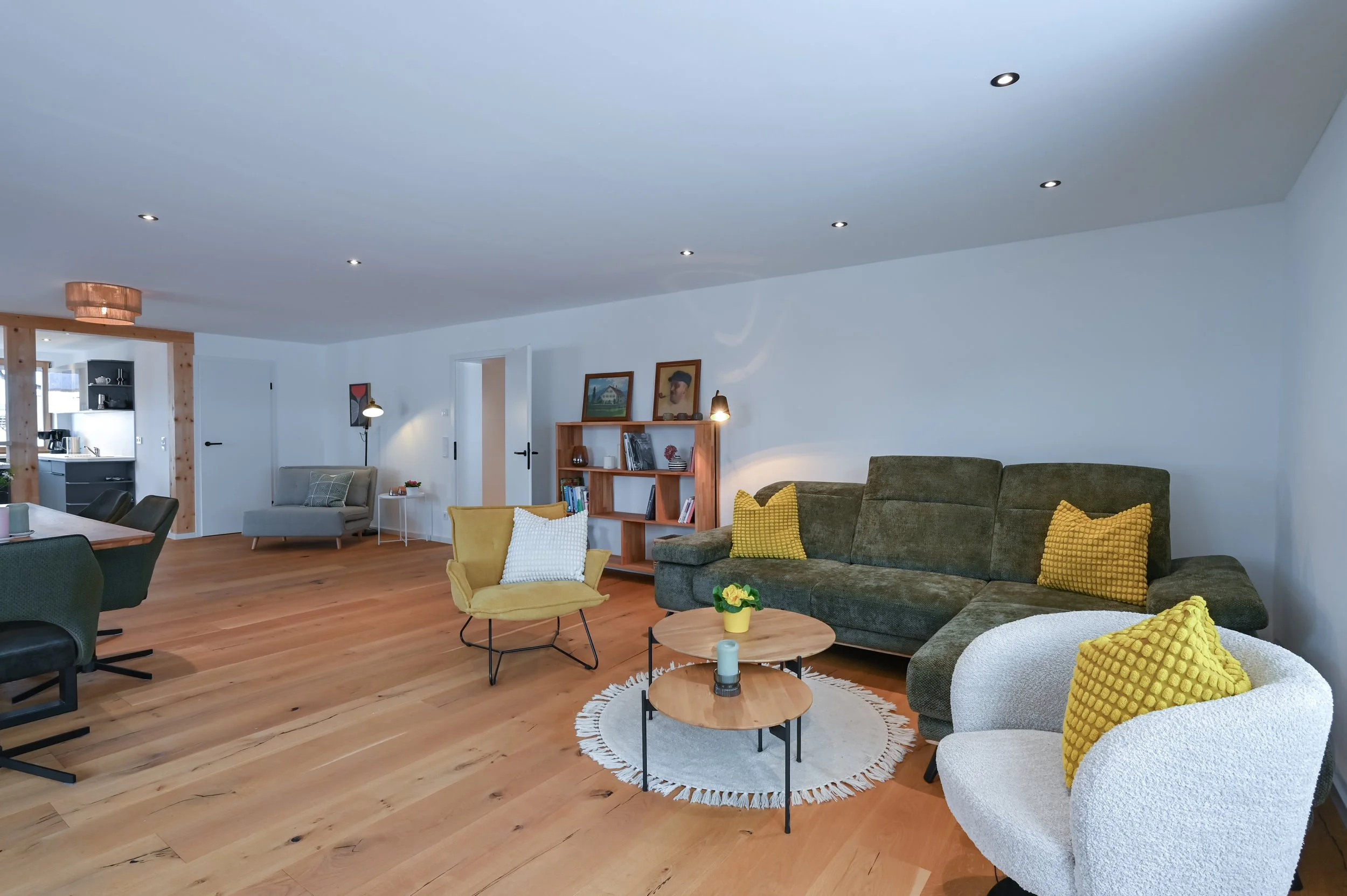 Ein modernes Wohnzimmer mit Holzfußboden, grauem Sofa mit gelben Kissen, weißen und gelben Sessel, kleinen Couchtischen, einem Bücherregal, mehreren Wandlampen und offenen Küchenbereich im Hintergrund.