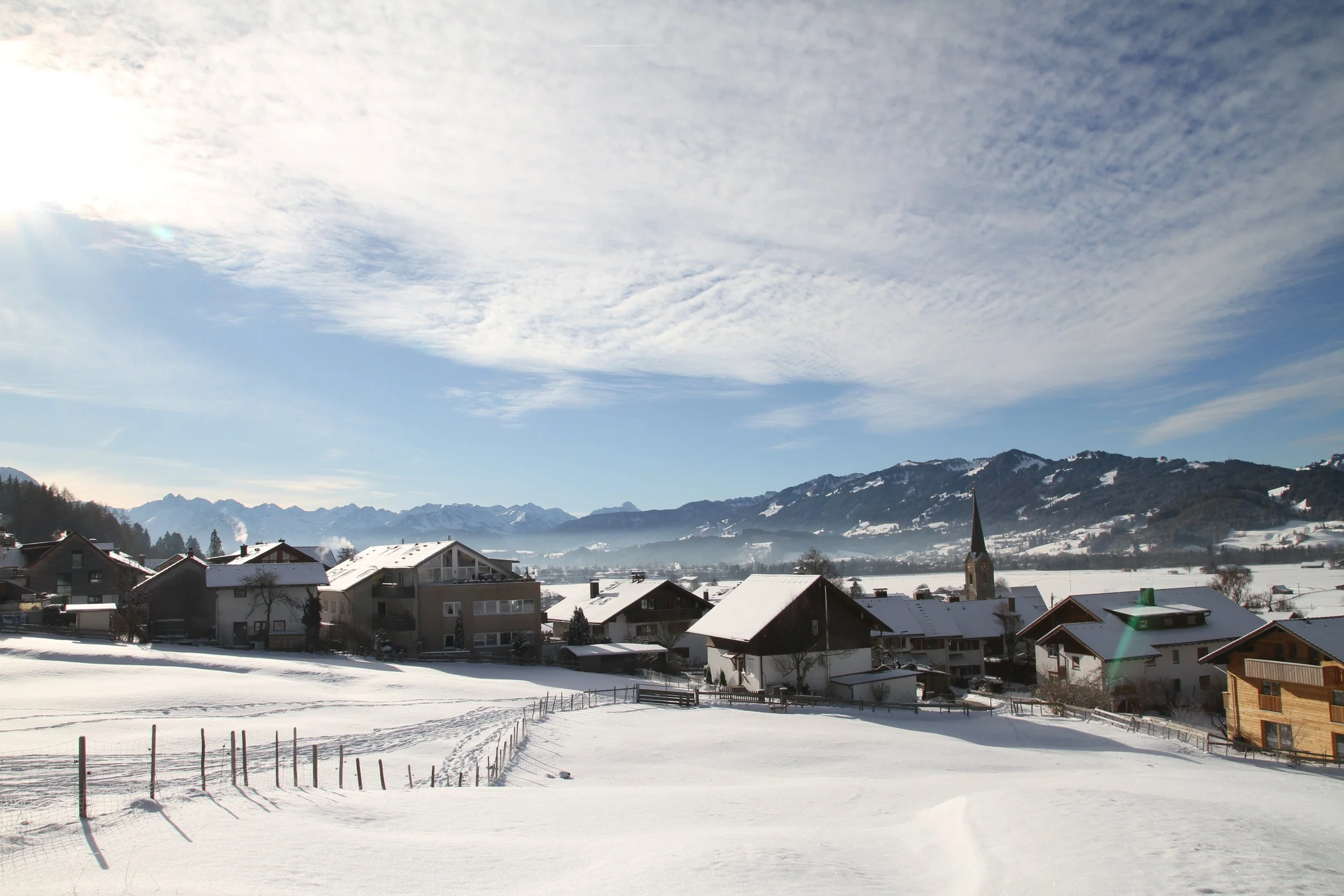 Schneebedeckte Häuser in einem Dorf vor einer Bergkulisse bei Sonnenlicht, mit einem klaren blauen Himmel und einigen Wolken.