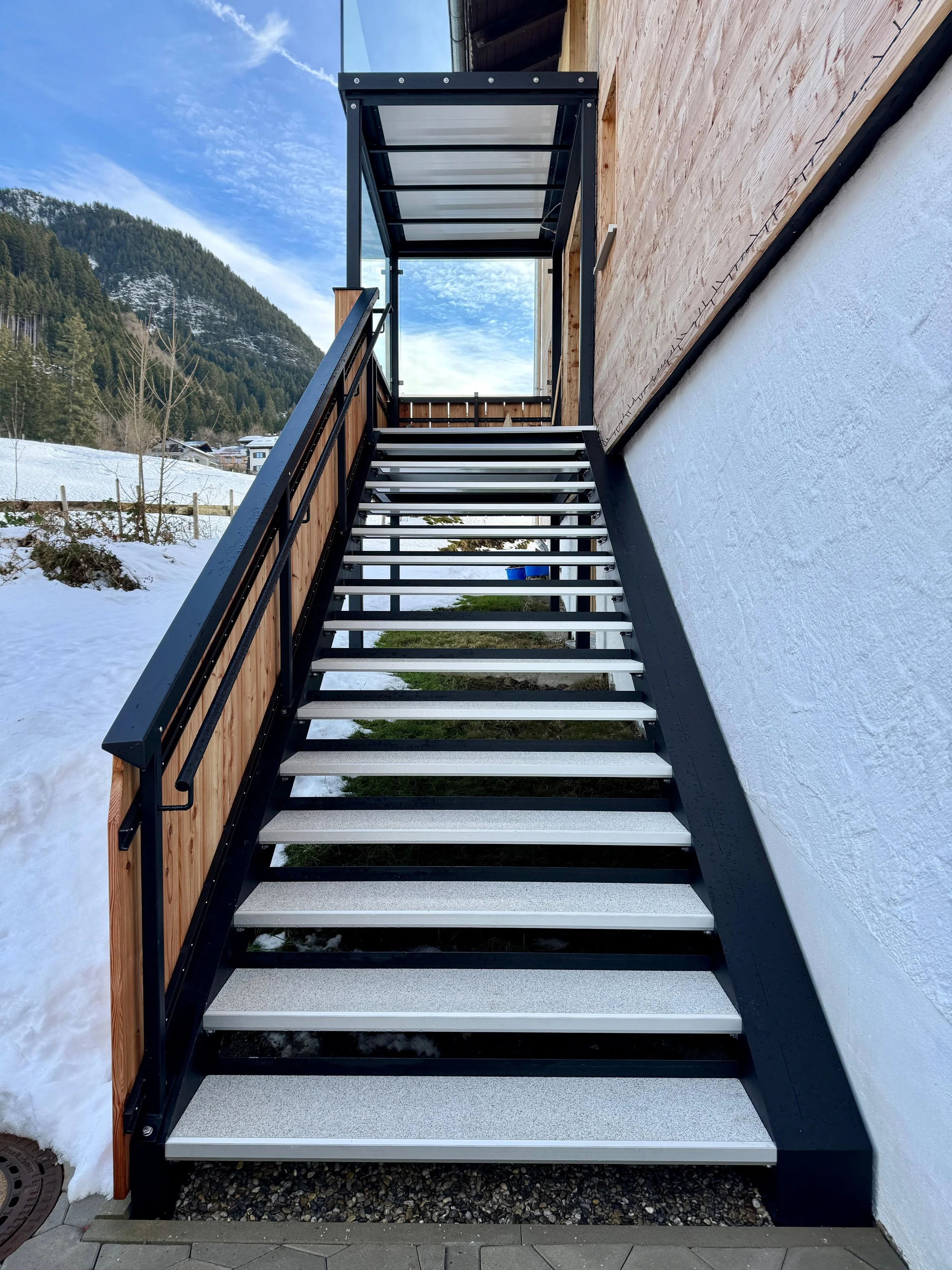 Außentreppe mit Metallstufen und schwarzem Geländer vor einem Haus, umgeben von Schnee und mit Berg im Hintergrund.
