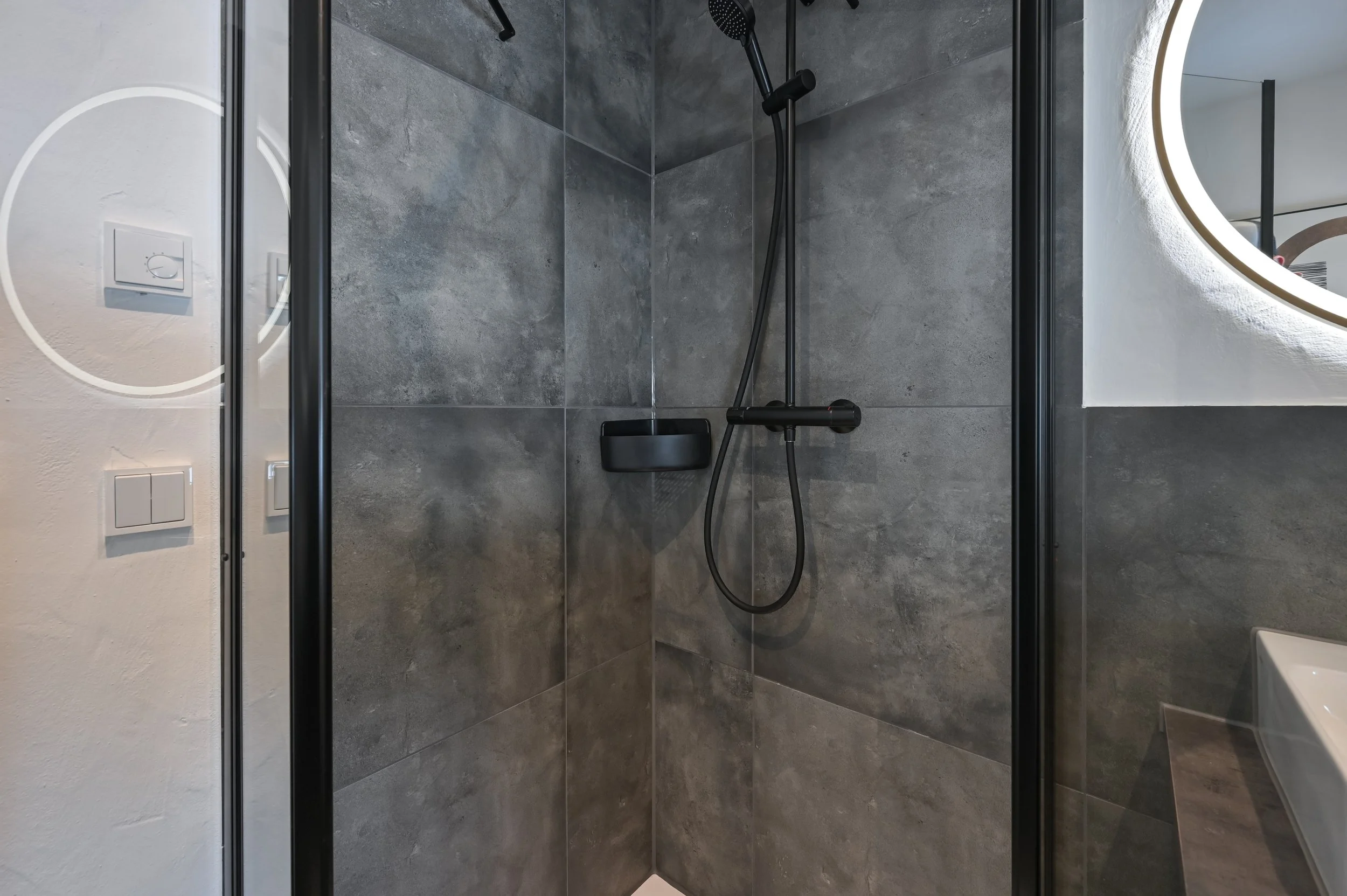 Moderner Duschraum mit grauen Fliesen, schwarzer Duschkopf und einem Spiegel mit LED-Beleuchtung.