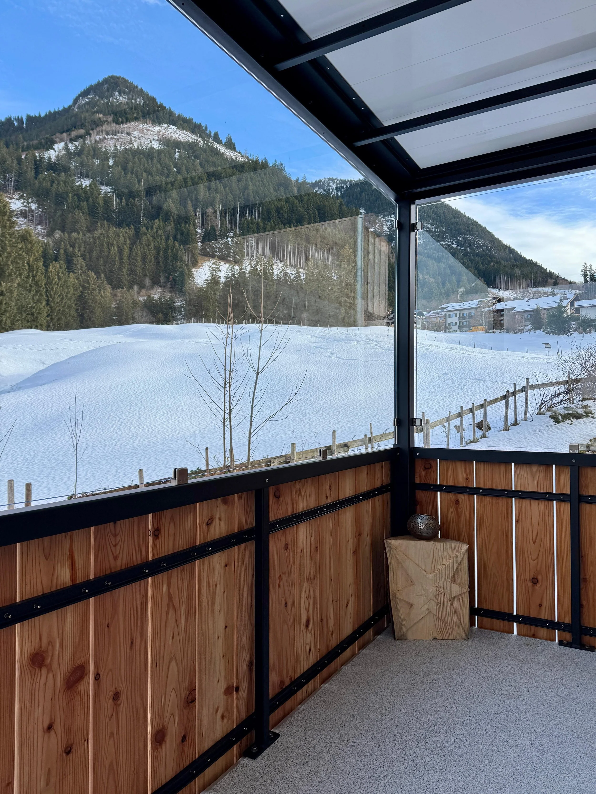 Blick von einer verglasten Veranda mit Holzverkleidung auf verschneite Hänge, Bäume und Berge in der Ferne