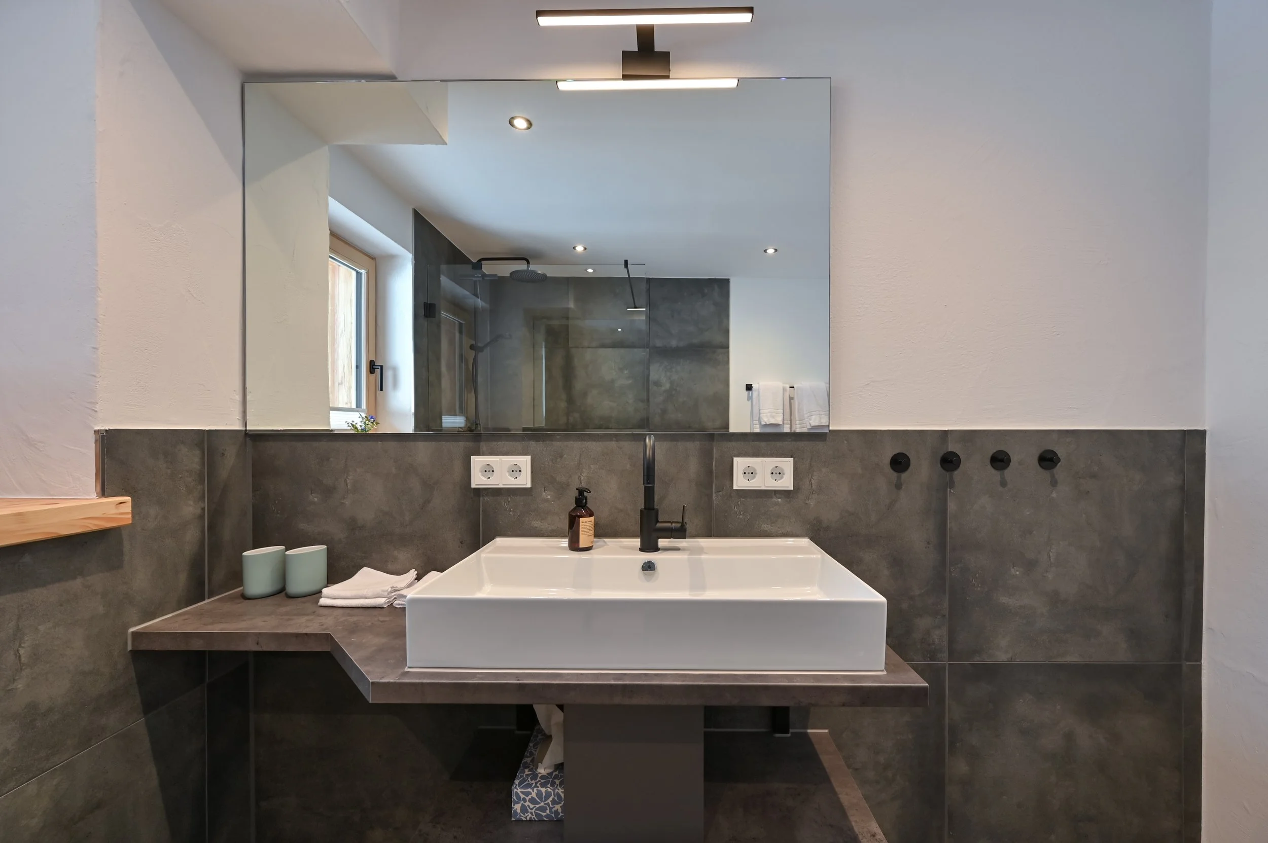 Moderner Waschbecken mit schwarzen Armaturen in einem Badezimmer. Hinter einem großen Spiegel sind Düsen für den Wasserhahn zu sehen.