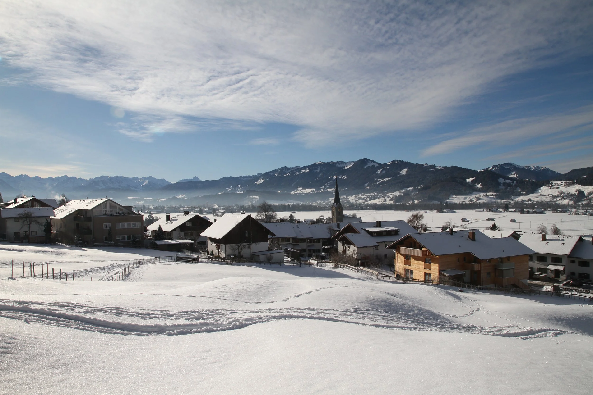 Schneebedecktes Dorf mit Häusern und Kirchturm vor Bergkulisse an einem klaren Wintertag.