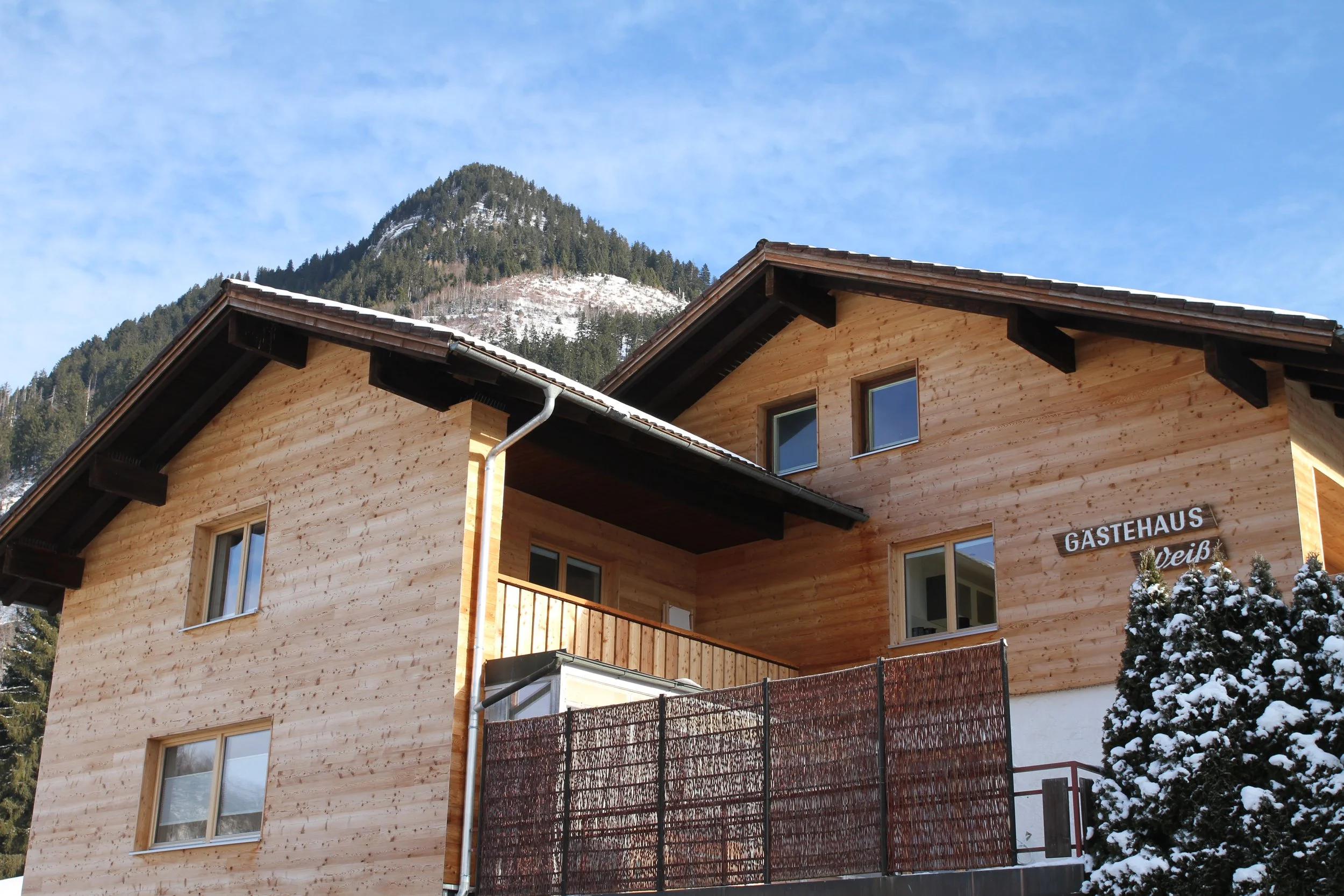 Holzhaus mit Balkon im Schnee, Berge im Hintergrund, bewölkter Himmel.
