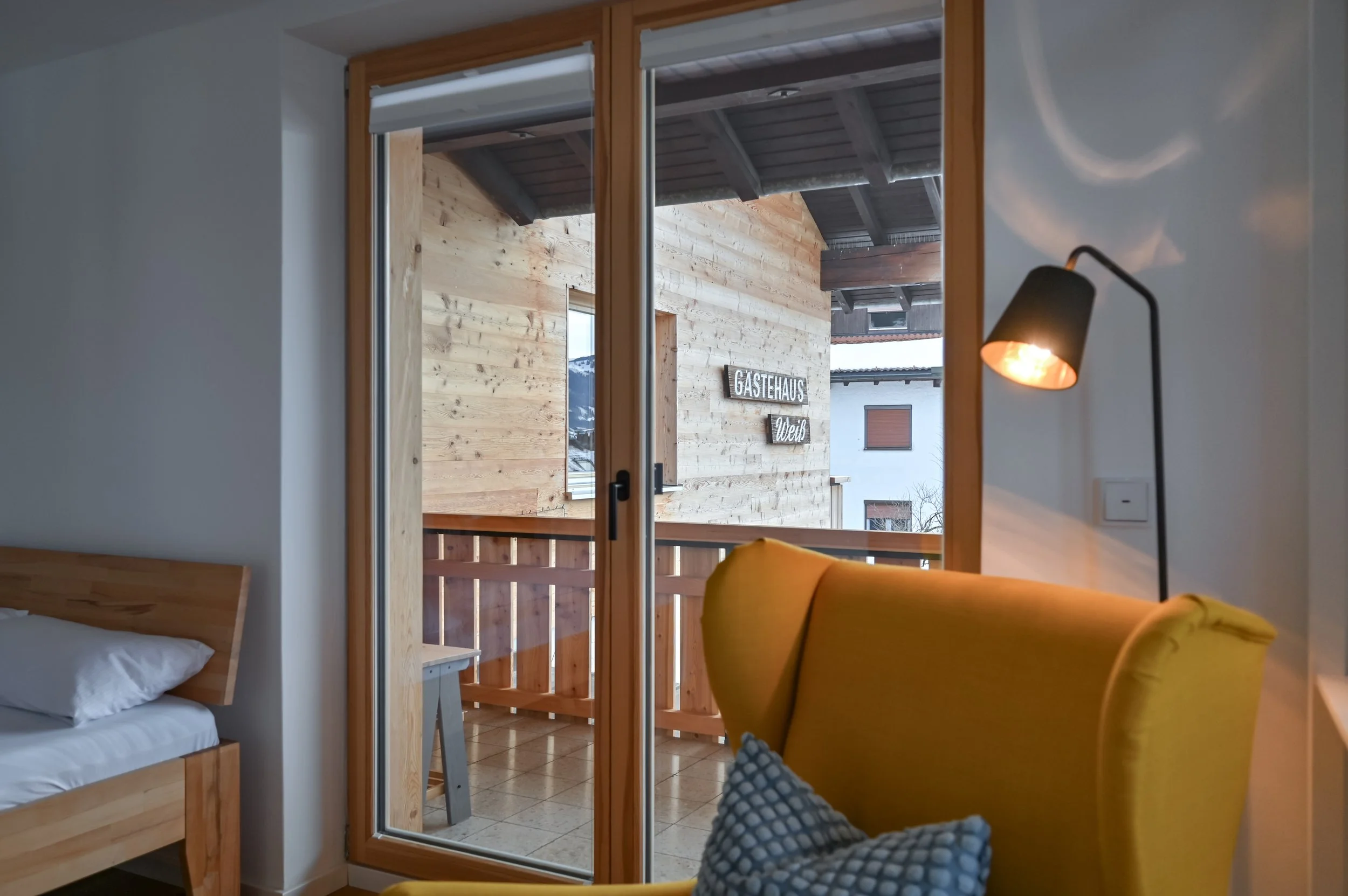 Innenraum eines Zimmers mit einem Fenster, einem gelben Sessel mit Kissen, einem Bett und einem Stehlampe, Blick auf einen Balkon mit Holzverkleidung und einer Beschilderung, die 'Gästehaus' und 'Weib' zeigt.